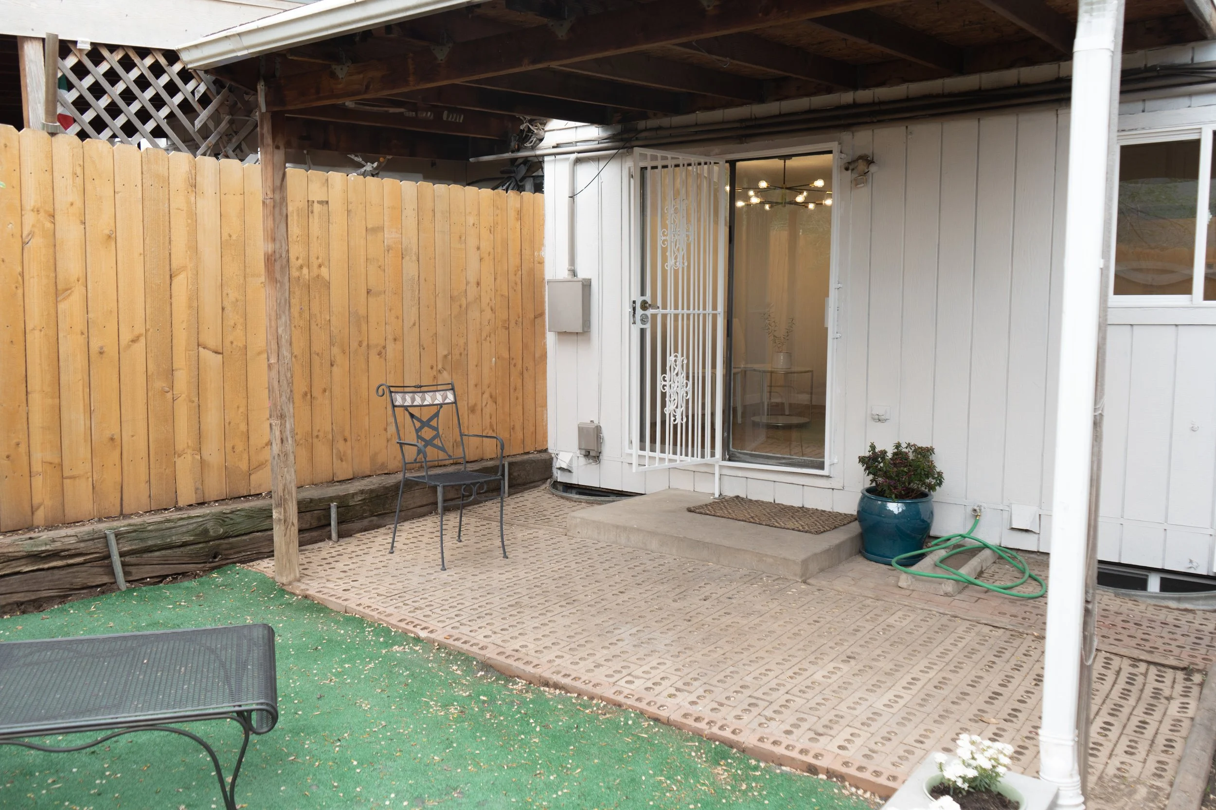 Small backyard patio with a wooden fence on one side and a white house wall on the other. There are metal chairs, a small table inside visible through a screen door, and decorative string lights. A green garden hose is on the ground, and there is a p