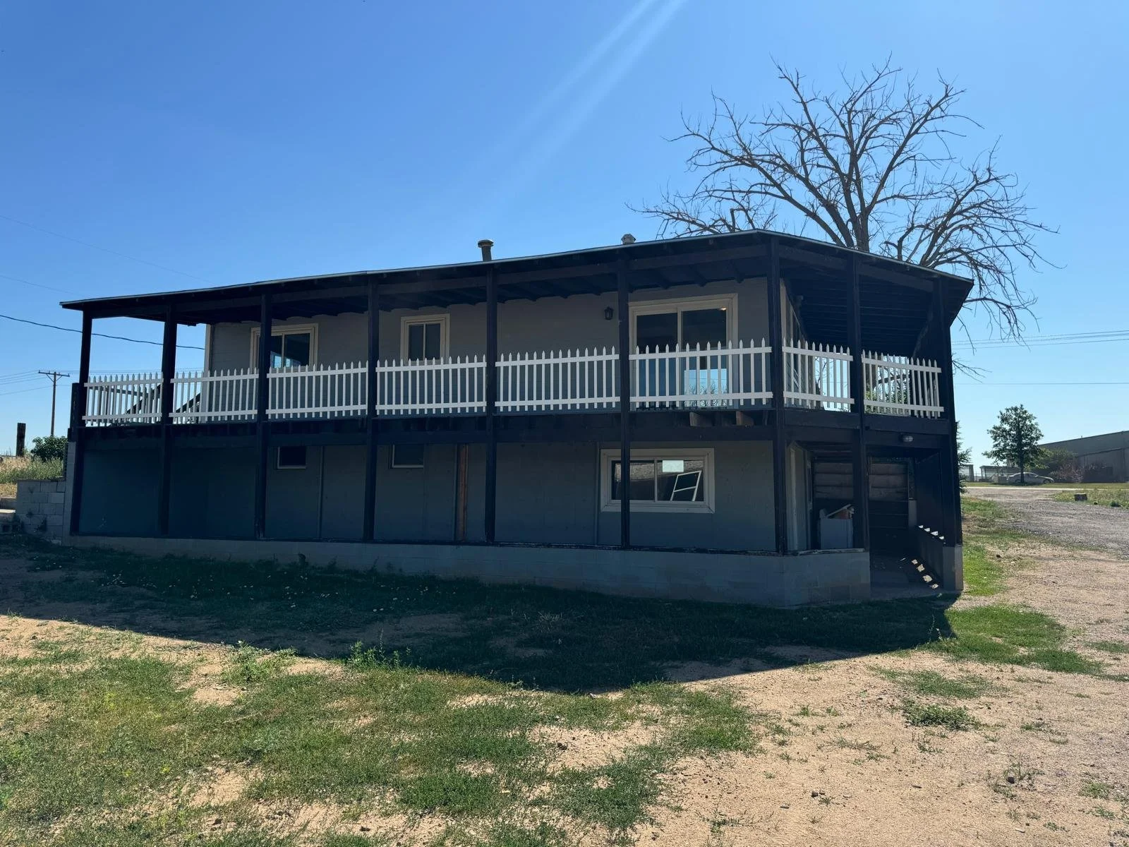A two-story house with a black frame, gray exterior walls, a wooden staircase, and a white picket fence along the upper balcony. There is a large, leafless tree behind the house and a bright blue sky.