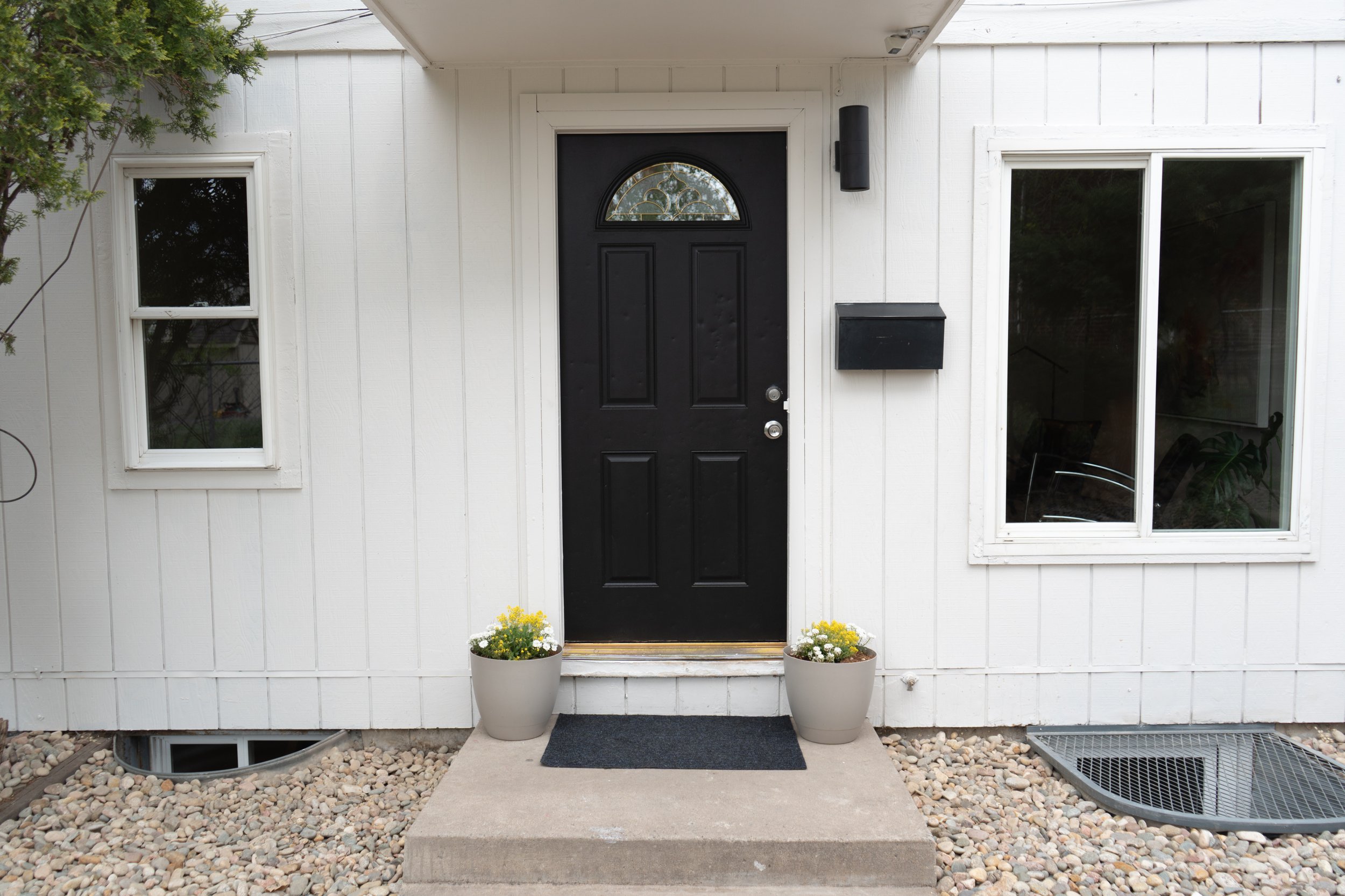 Front entrance of a house with a black door, two windows, a black mailbox, a black wall-mounted light, and two potted plants with yellow and white flowers on either side of the door. Pebble ground and a small concrete porch.