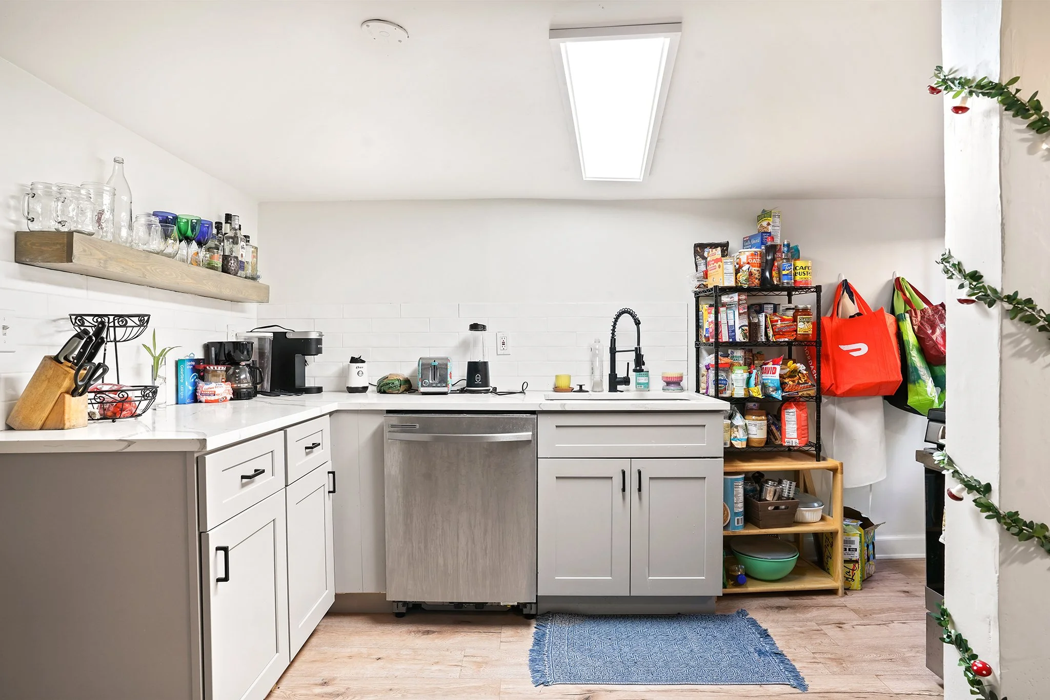 A kitchen with white cabinets, a black rack with snack bags, a wooden shelf, and various kitchen appliances on the counter. There is a window in the ceiling and a green plant draped over the corner.