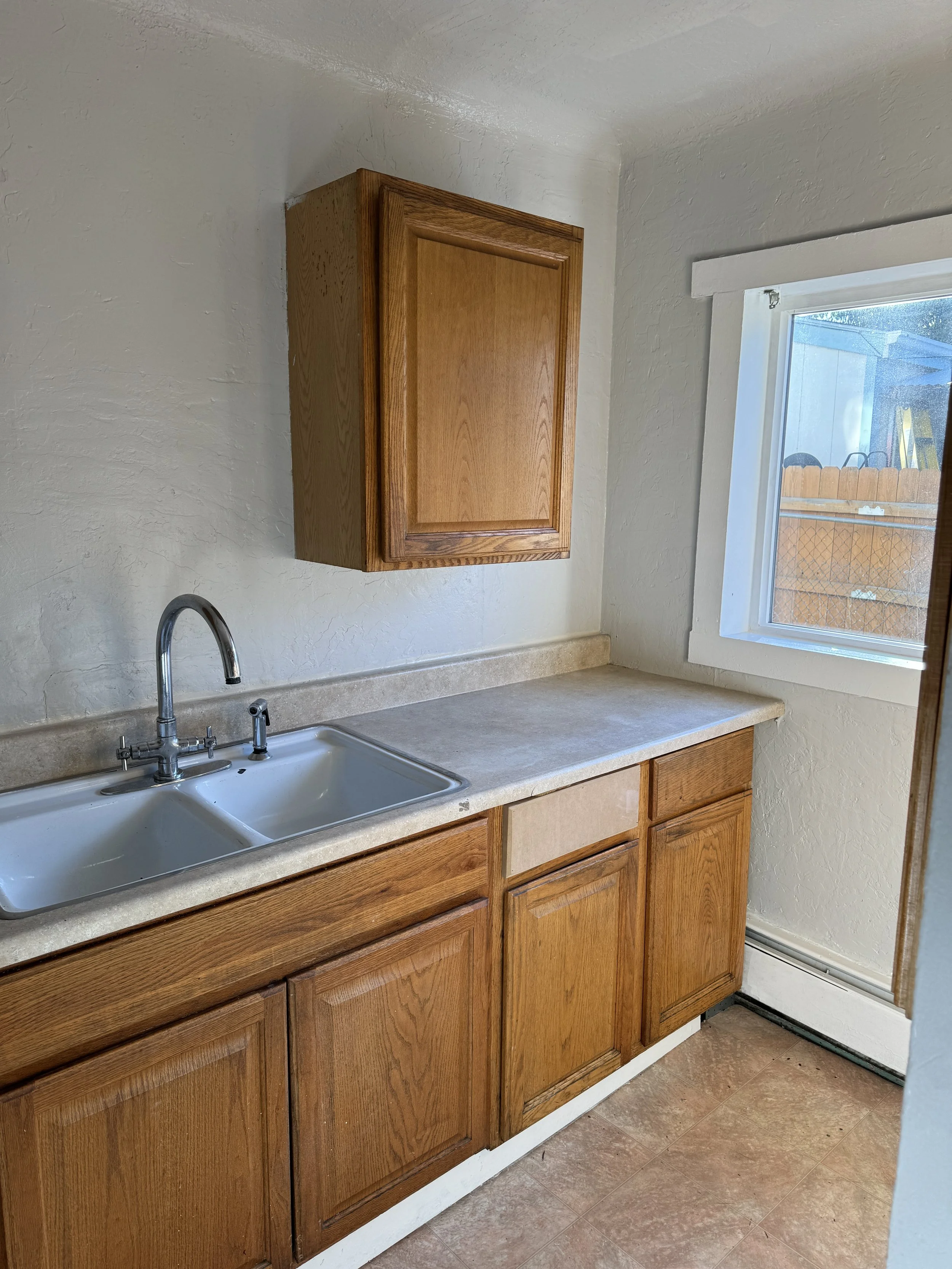 A small kitchen corner with a wooden cabinet above and below the beige countertop, a double stainless steel sink with a faucet, and a window with a view of a wooden fence and outdoor yard.