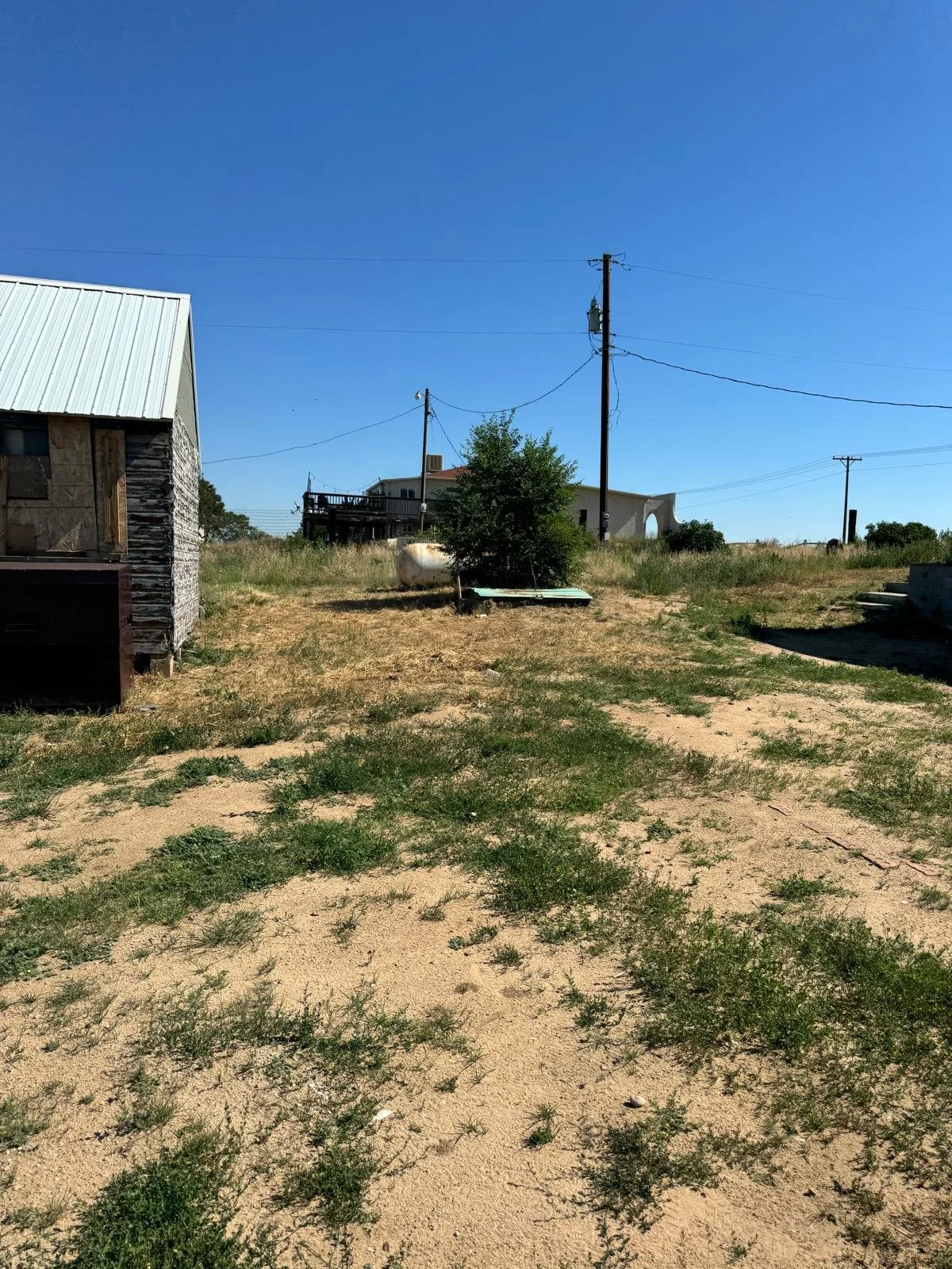 A rural scene with a sandy and grassy yard, a weathered wooden shed, power lines, a tree, and a few houses in the background under a clear blue sky.