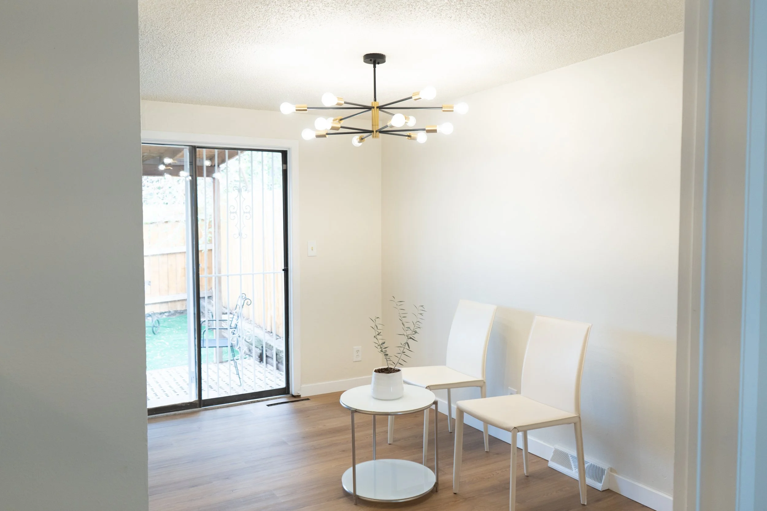 A bright dining area with a sliding glass door leading to an outdoor patio, two white chairs, a small round white table with a potted plant, a modern black and gold chandelier, and hardwood flooring.