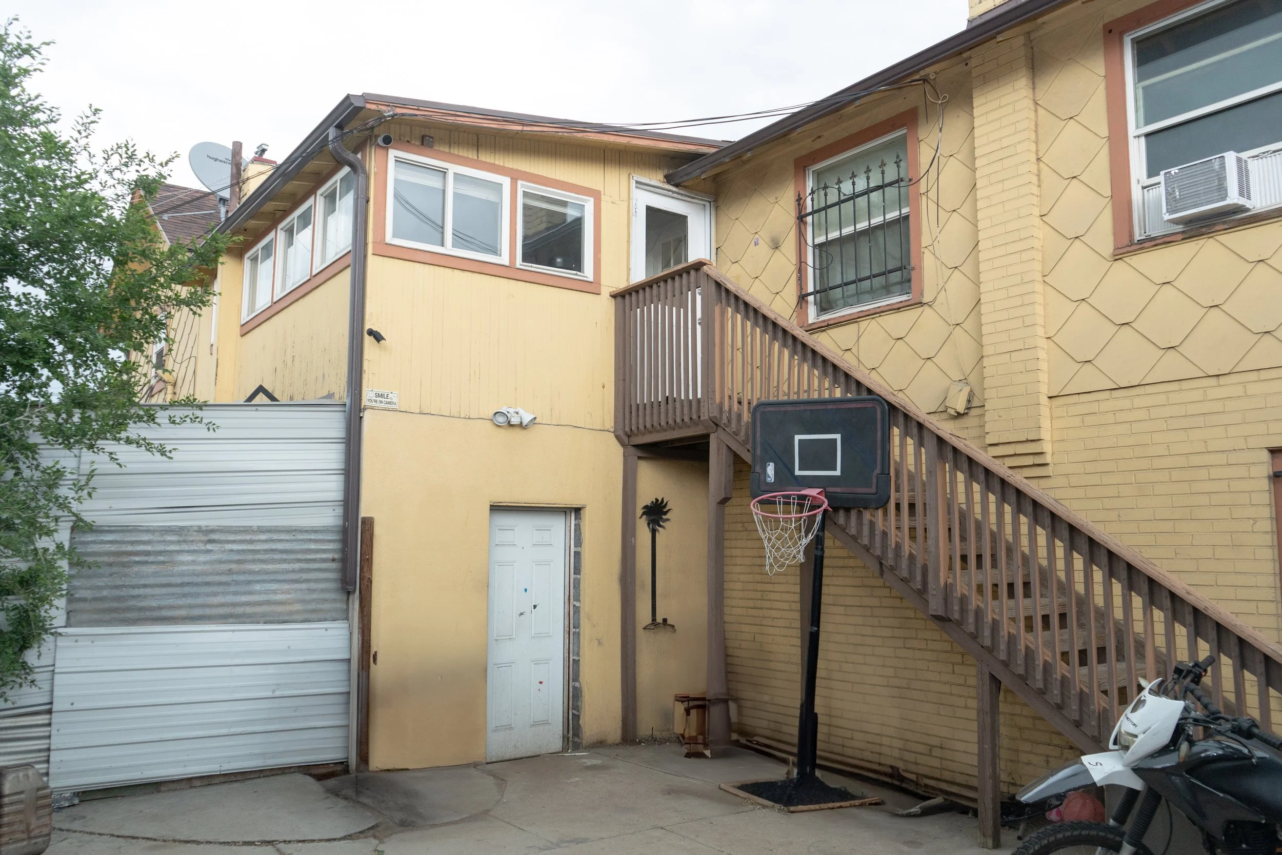 Backyard of a yellow house with a basketball hoop set up, a motorcycle in the corner, and a wooden staircase leading to a door and windows on the second floor.