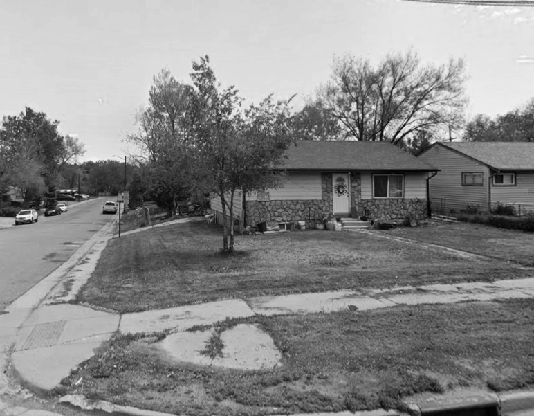 A small house with a stone and yellow exterior, front yard with grass and a tree, sidewalk, and neighboring houses on a residential street.
