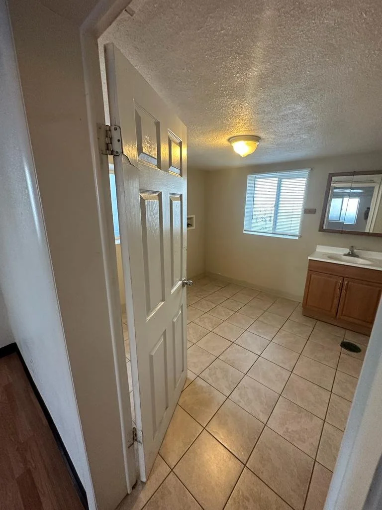 Empty bathroom with tiled floor, a window with blinds, a small sink with wooden cabinet, and a mirror above the sink.