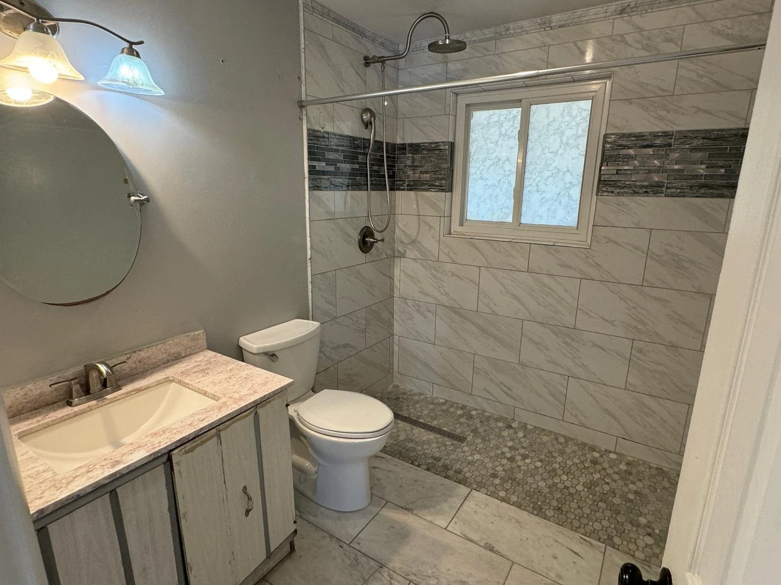 Bathroom with a vanity, toilet, and walk-in shower with stone tile accents, a small window, and a mirror with three wall-mounted lights.