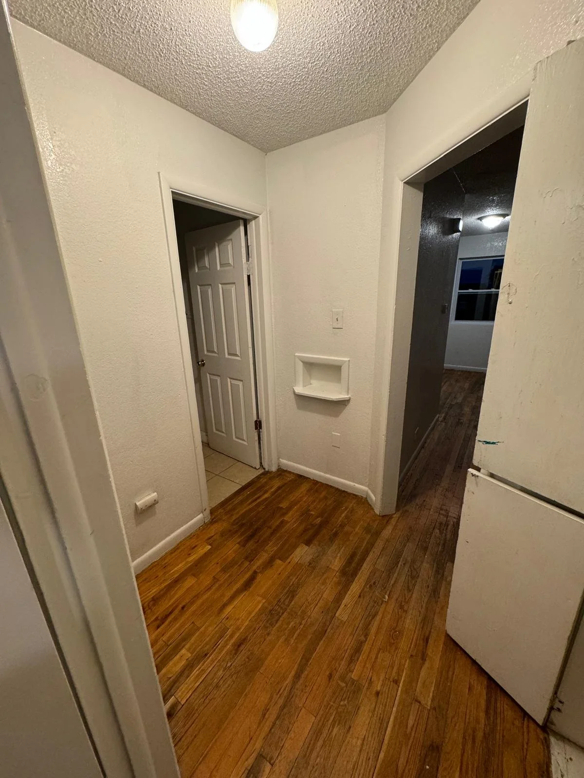 A small hallway with white textured walls, a wooden floor, a closed six-panel door, and an open doorway leading to another room with a window and dark walls. A white shelf is built into the wall near the open doorway.