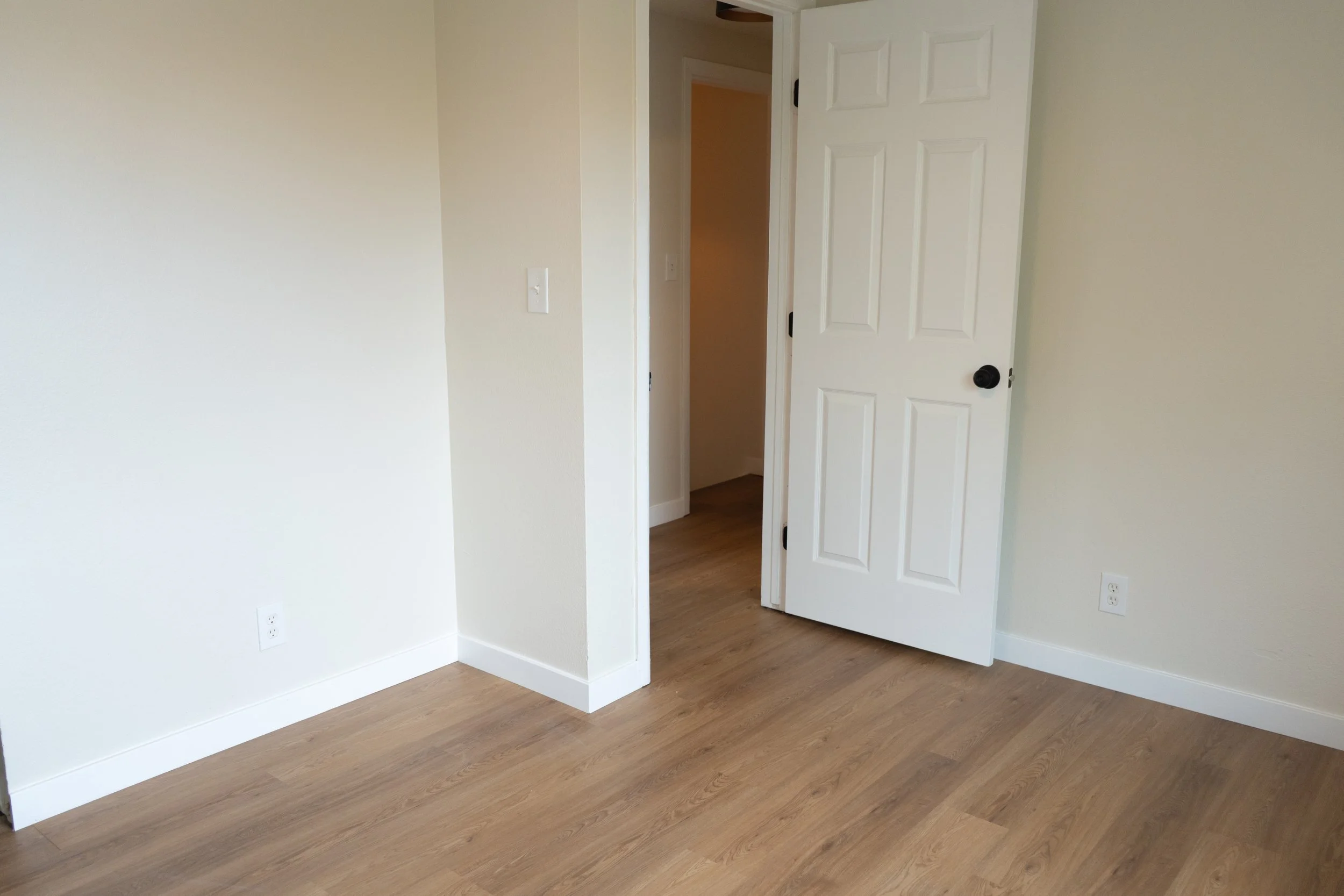 Empty room with light wood flooring, white walls, an open white door, and electrical outlets.