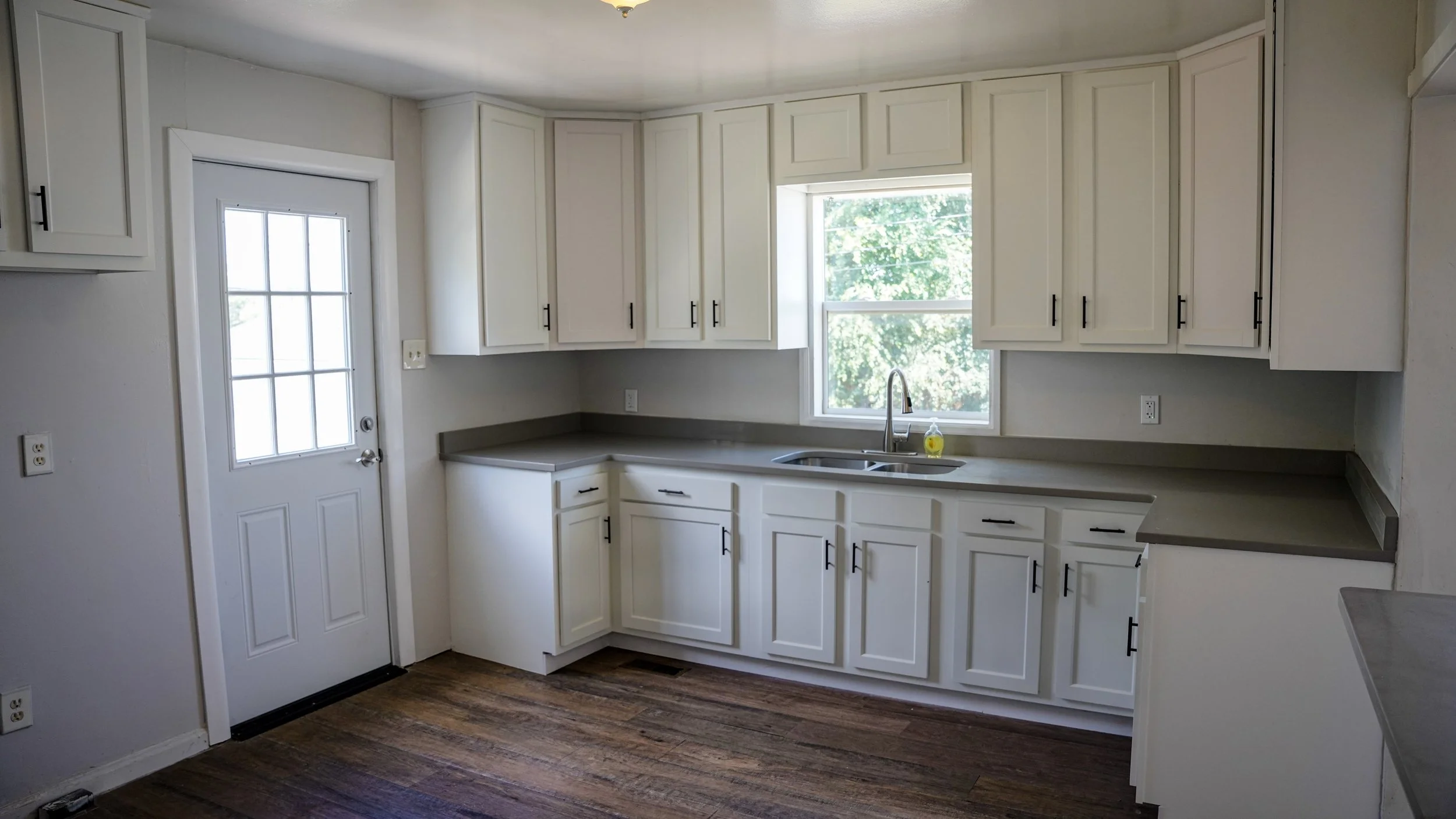 A kitchen with white cabinets, gray countertops, a stainless steel sink, a window above the sink, and a door with a window on the left. The floor is wooden, and there are electrical outlets on the walls.