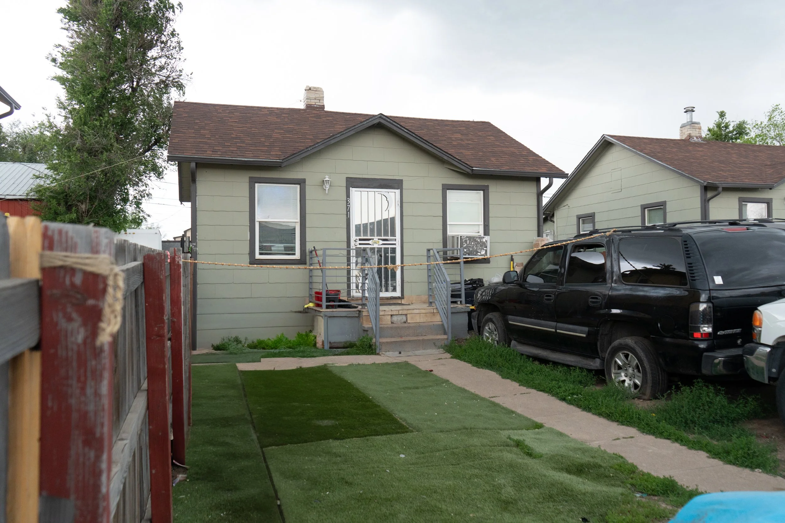 Backyard view of a green house with stairs leading to the door, black vehicle parked beside it, and a wooden fence on the left side.