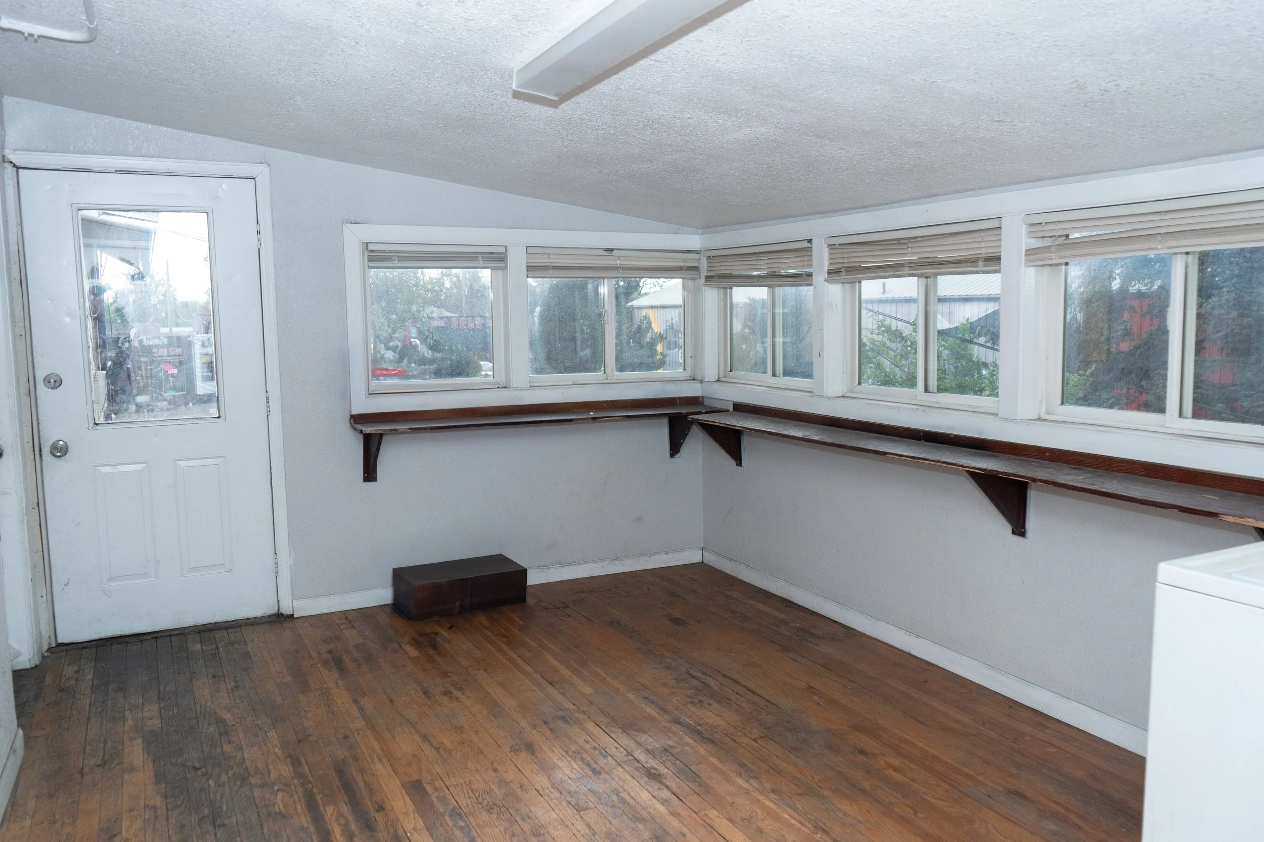 Empty room with hardwood floor, white walls, and multiple windows with blinds, featuring a door to the outside, a long wooden shelf on the wall, and a small black box on the floor.