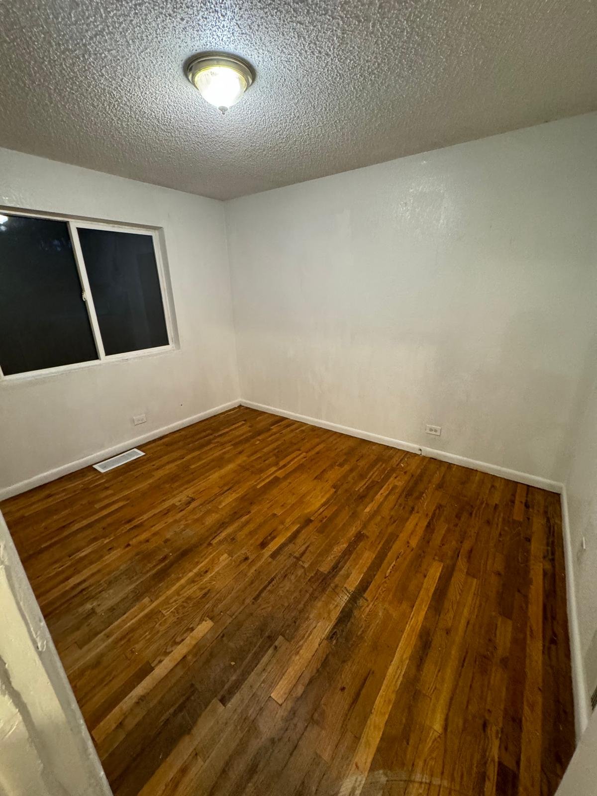 Empty room with white drywall walls, hardwood floor, a window on the left wall, and a ceiling light fixture.