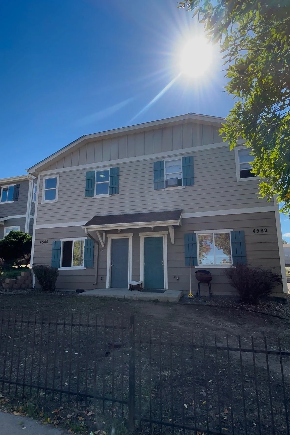 A two-story beige house with blue window shutters, two front doors, and a small porch, under a bright sun in a clear blue sky.