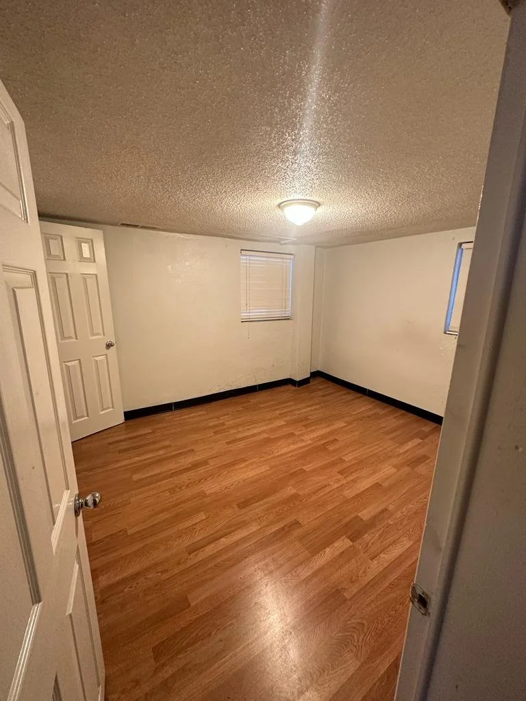 Empty room with white walls, wooden floor, small window with closed blinds, ceiling light, and door slightly open.