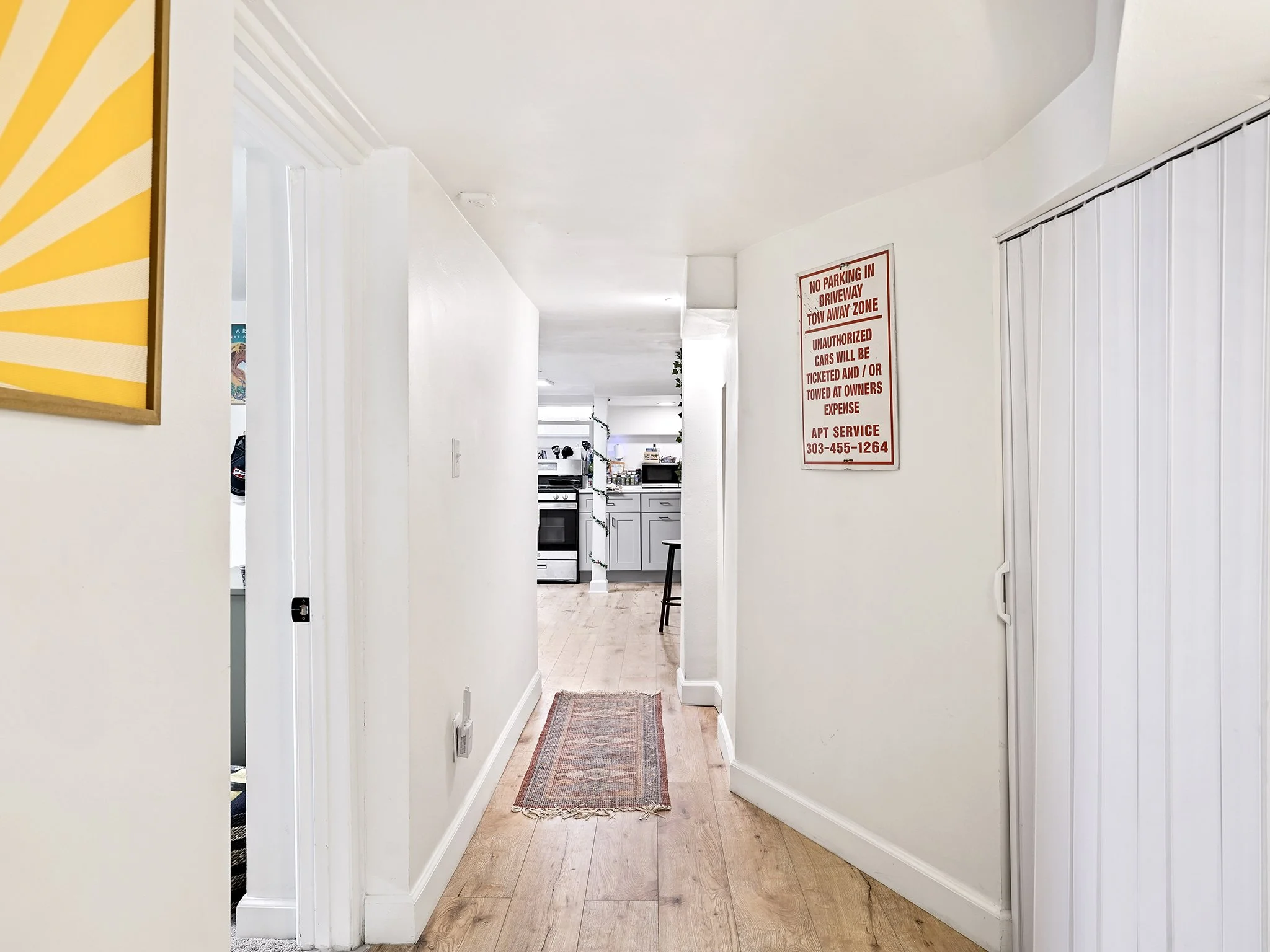 A hallway leading into a kitchen area with a stove and countertop, with a rug on the wooden floor and a white wall on the right side with a vertical blind window shade, and a red and white warning sign on the wall.