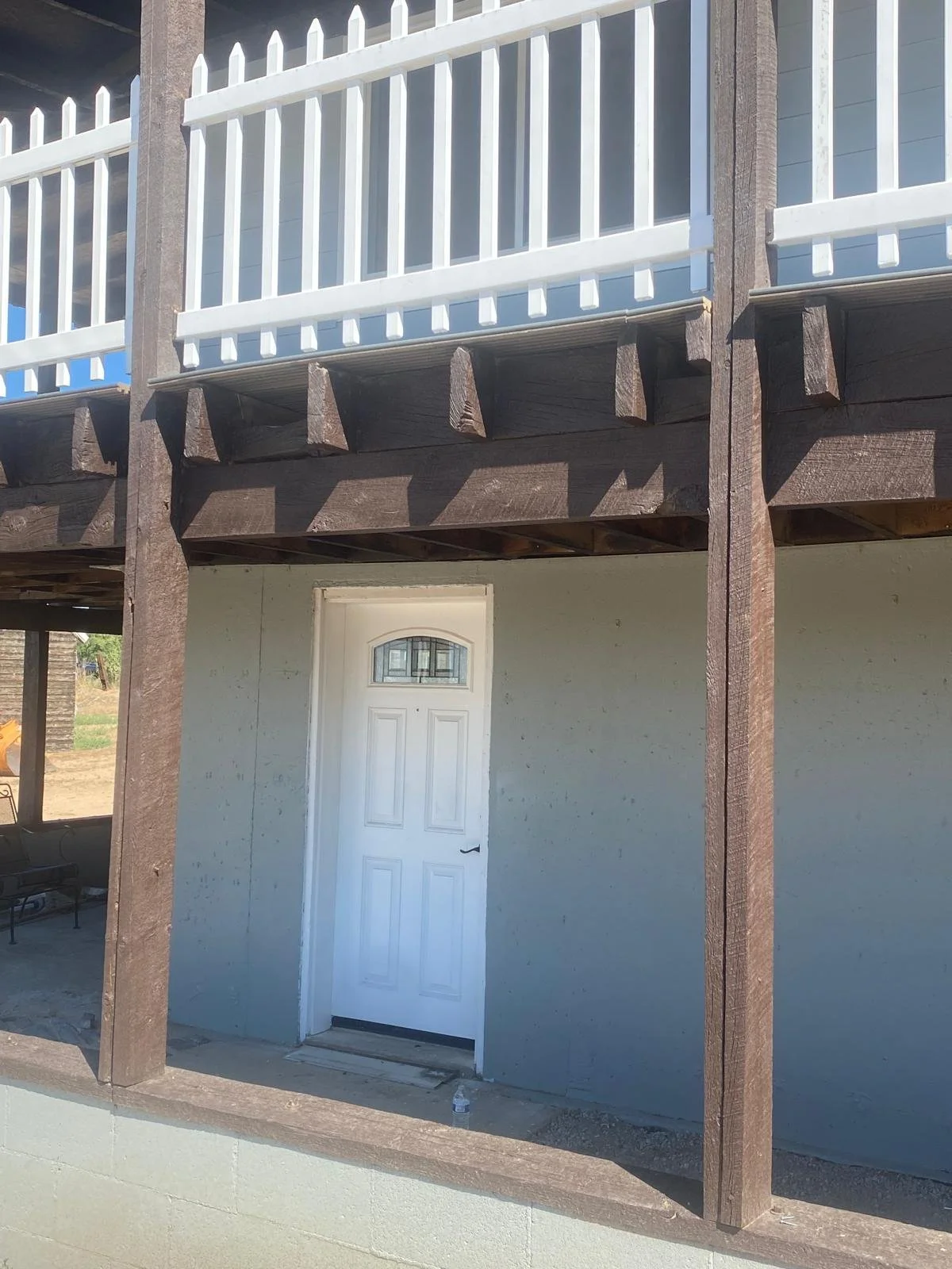 View of the underside of a balcony or deck with white railing and brown wooden supports, showing a white door on the ground level.