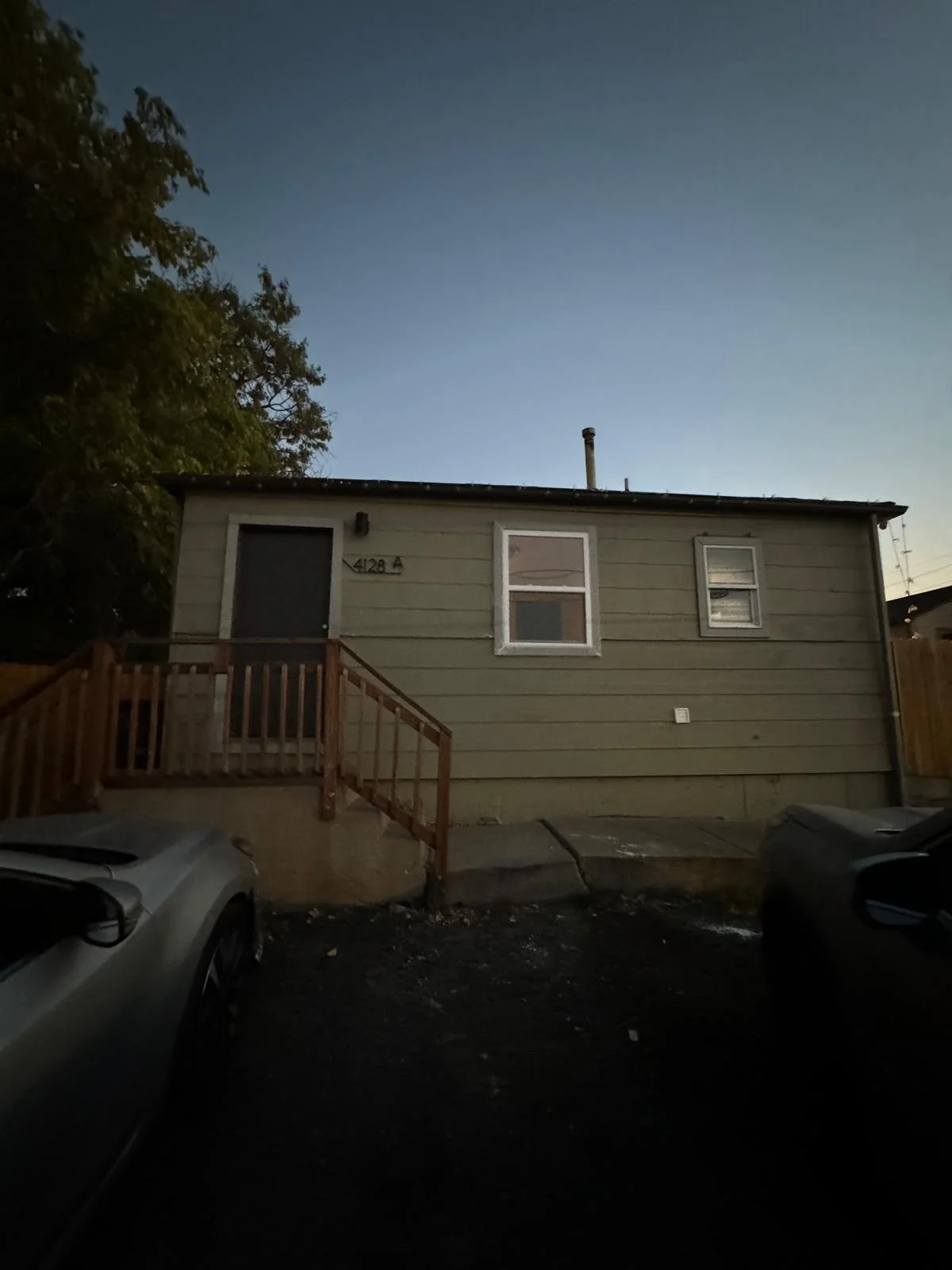 A small, two-story house with light green siding, two windows, and a small wooden staircase leading to a back door, seen during dusk. Two cars are parked in front, and a tree is visible on the left.