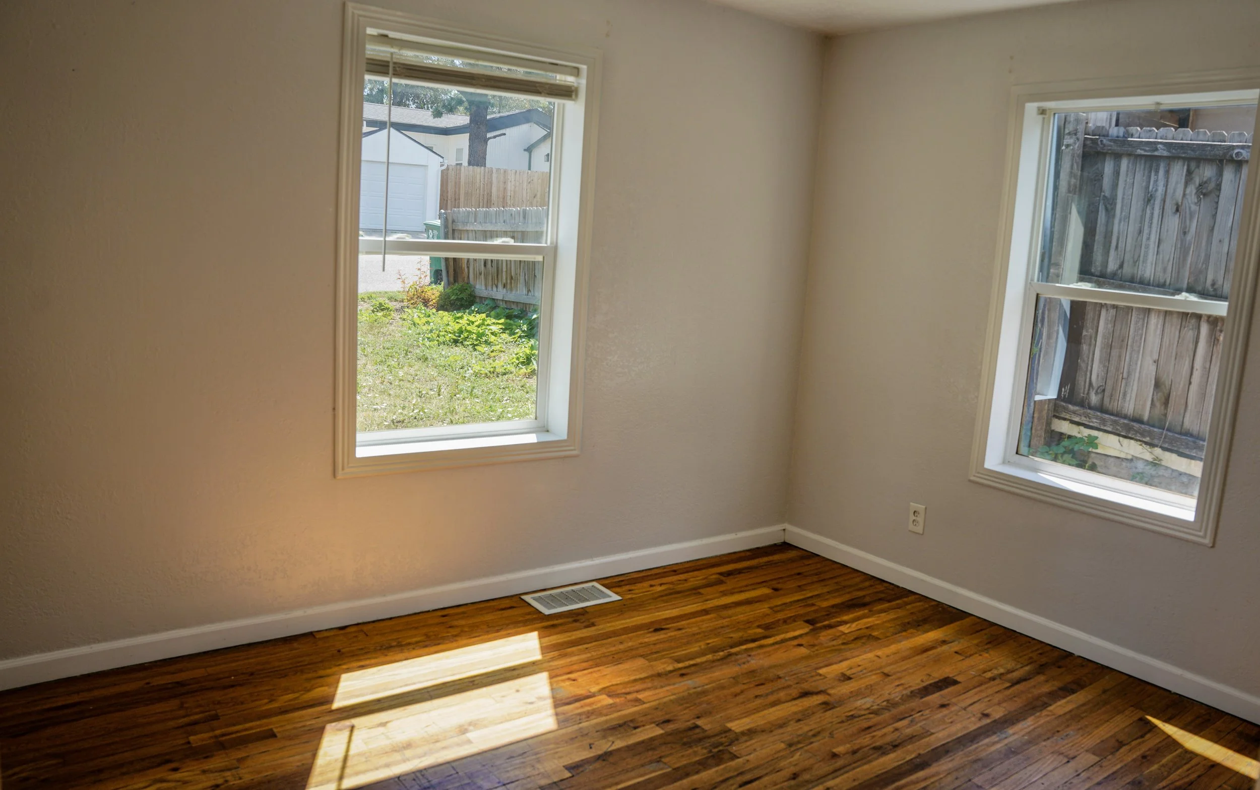 Empty room with hardwood floors, two windows showing a small yard with a fence, shed, and trees outside.