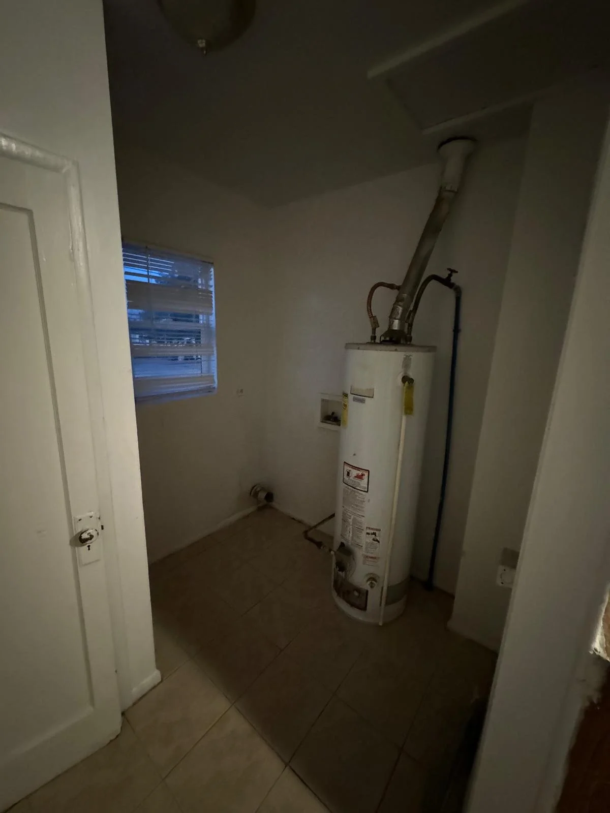 A small laundry or utility room with a white water heater, tiled floor, small window with blinds, and white walls.