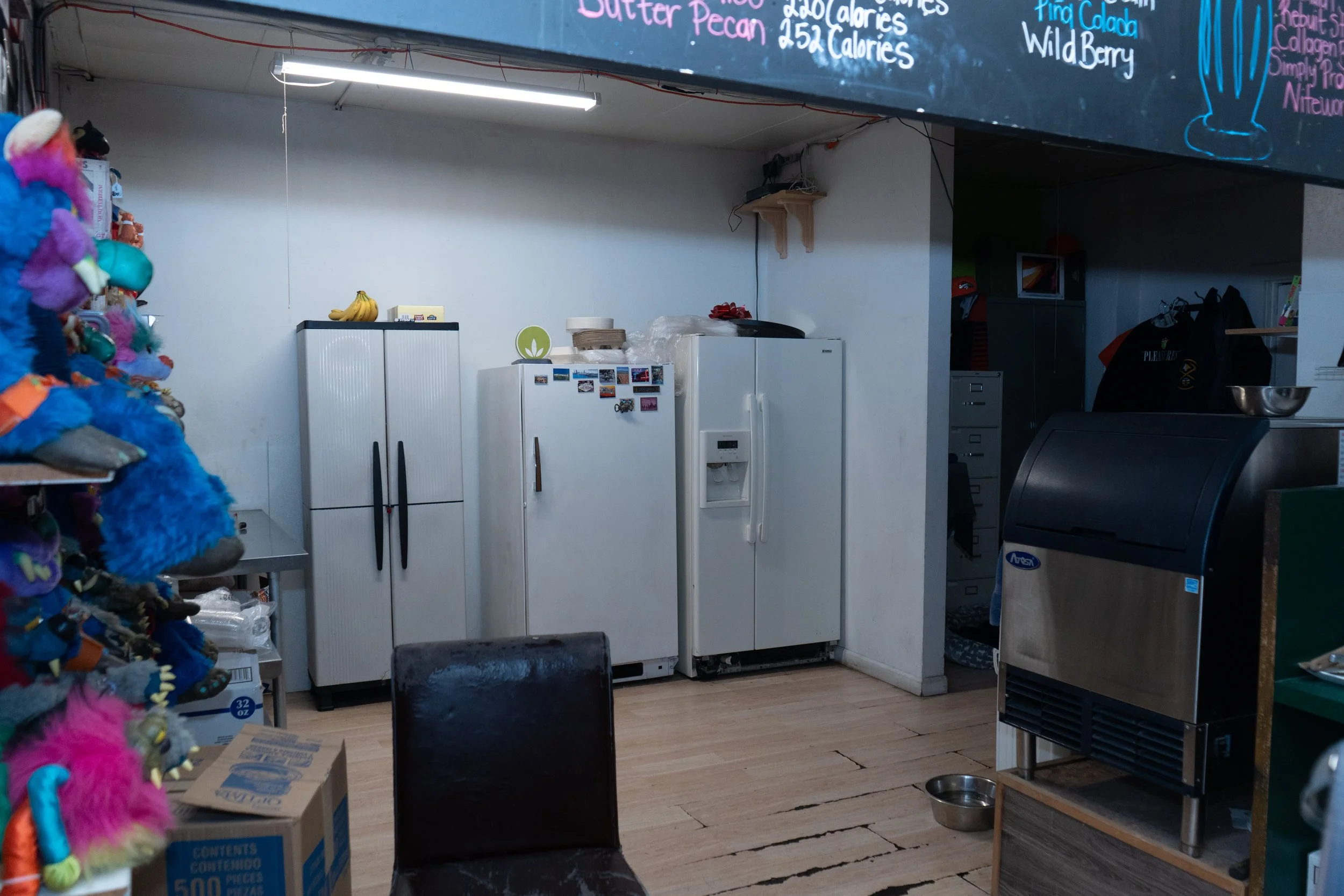 A cluttered room with a white fridge, a standing freezer, a white cabinet, and a few other appliances. Items such as bananas and a small decorative item are on top of the furniture. A black chair is in the foreground, and a snack machine is on the ri