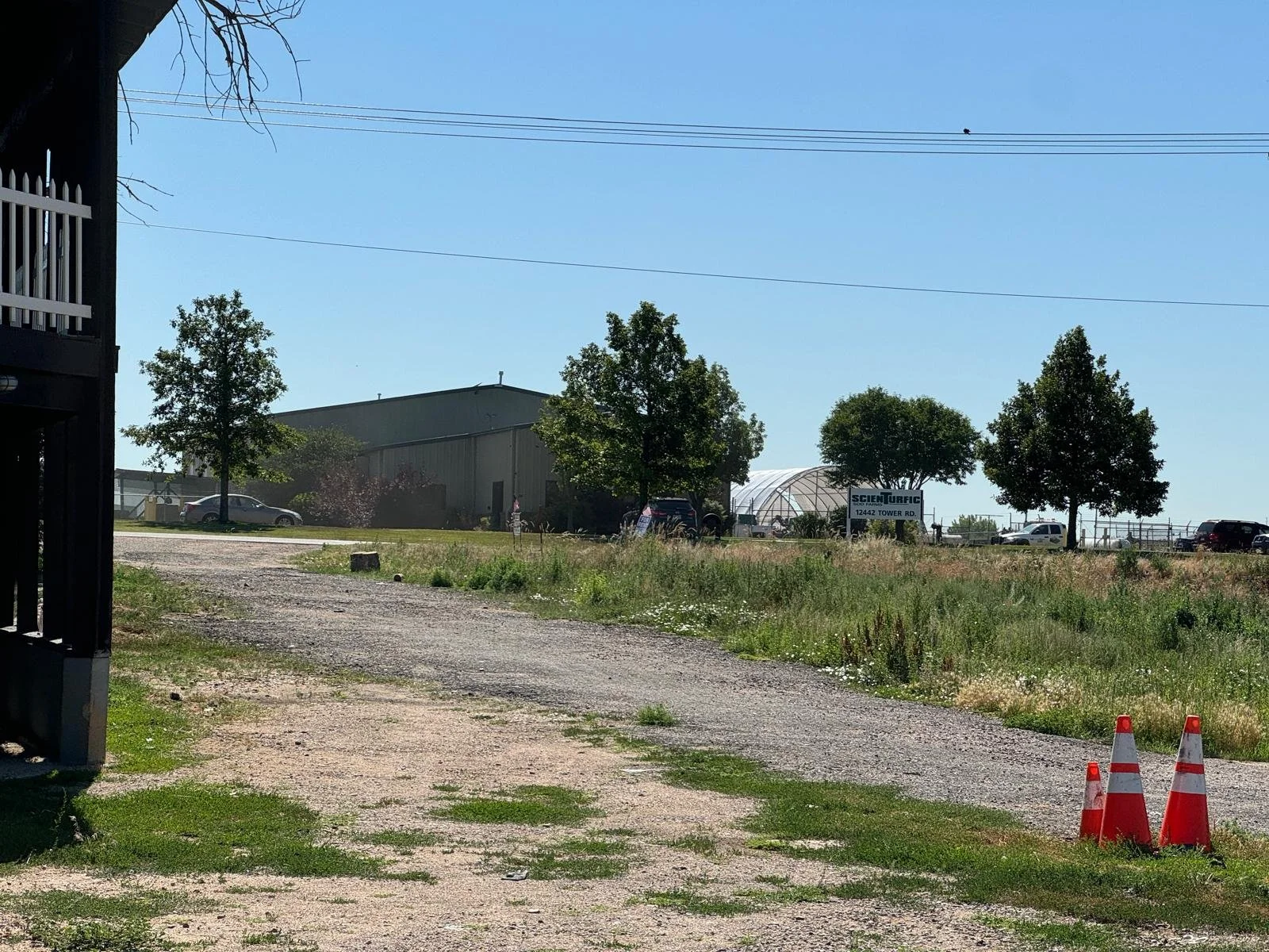 Grassy area with a gravel path, three orange traffic cones, several trees, a building, and parked cars in the background under a clear blue sky.