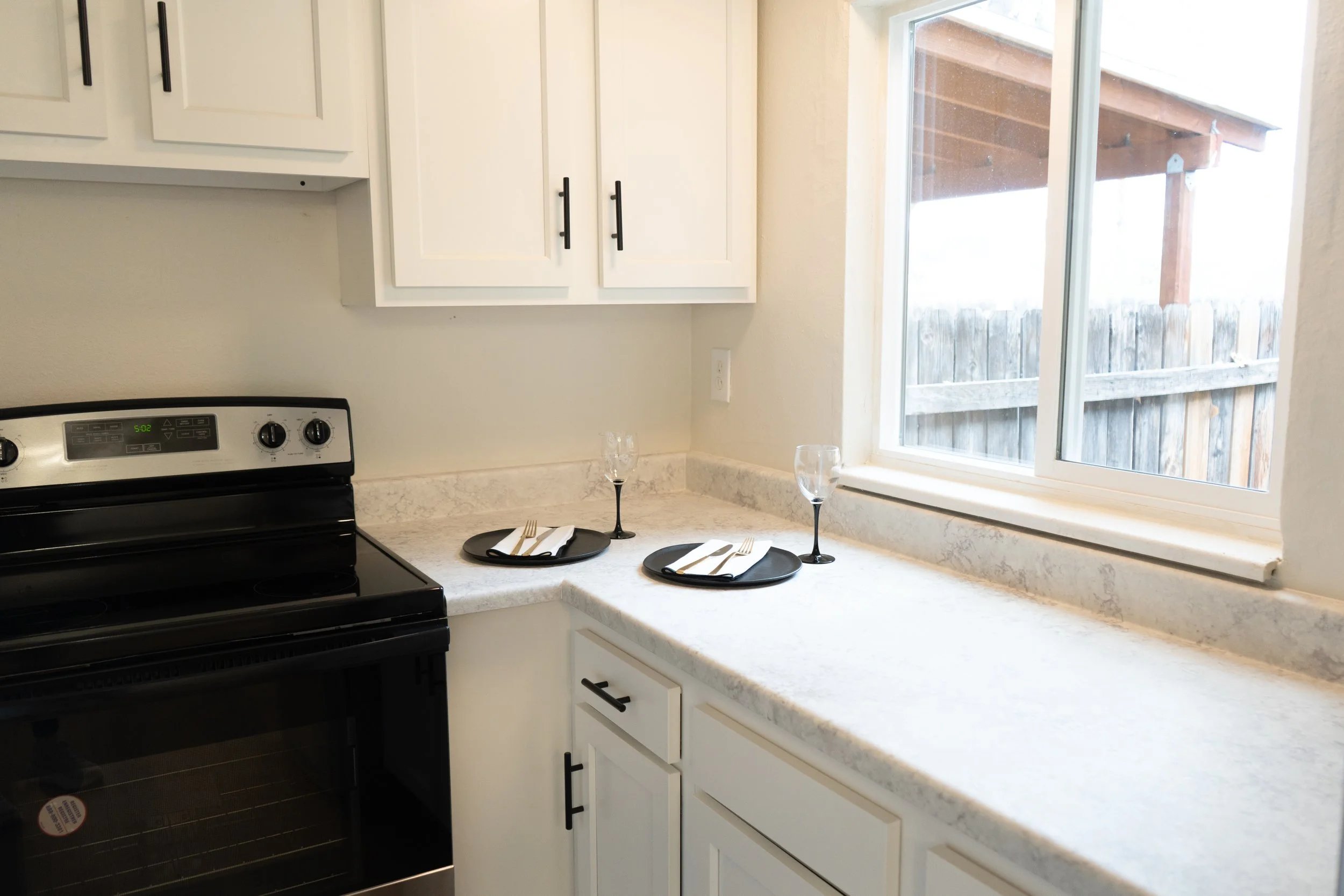 Kitchen counter with table setting for two, including black plates, napkins, cutlery, and empty wine glasses near a window with a wooden fence outside.