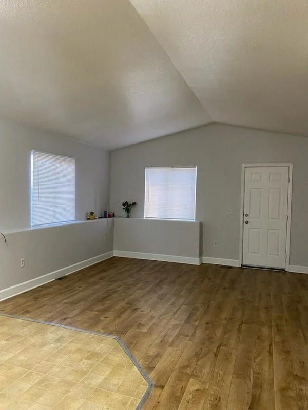 Empty room with hardwood floors, white walls, and two windows with blinds, one shelf with small items, a potted plant, and a white door.