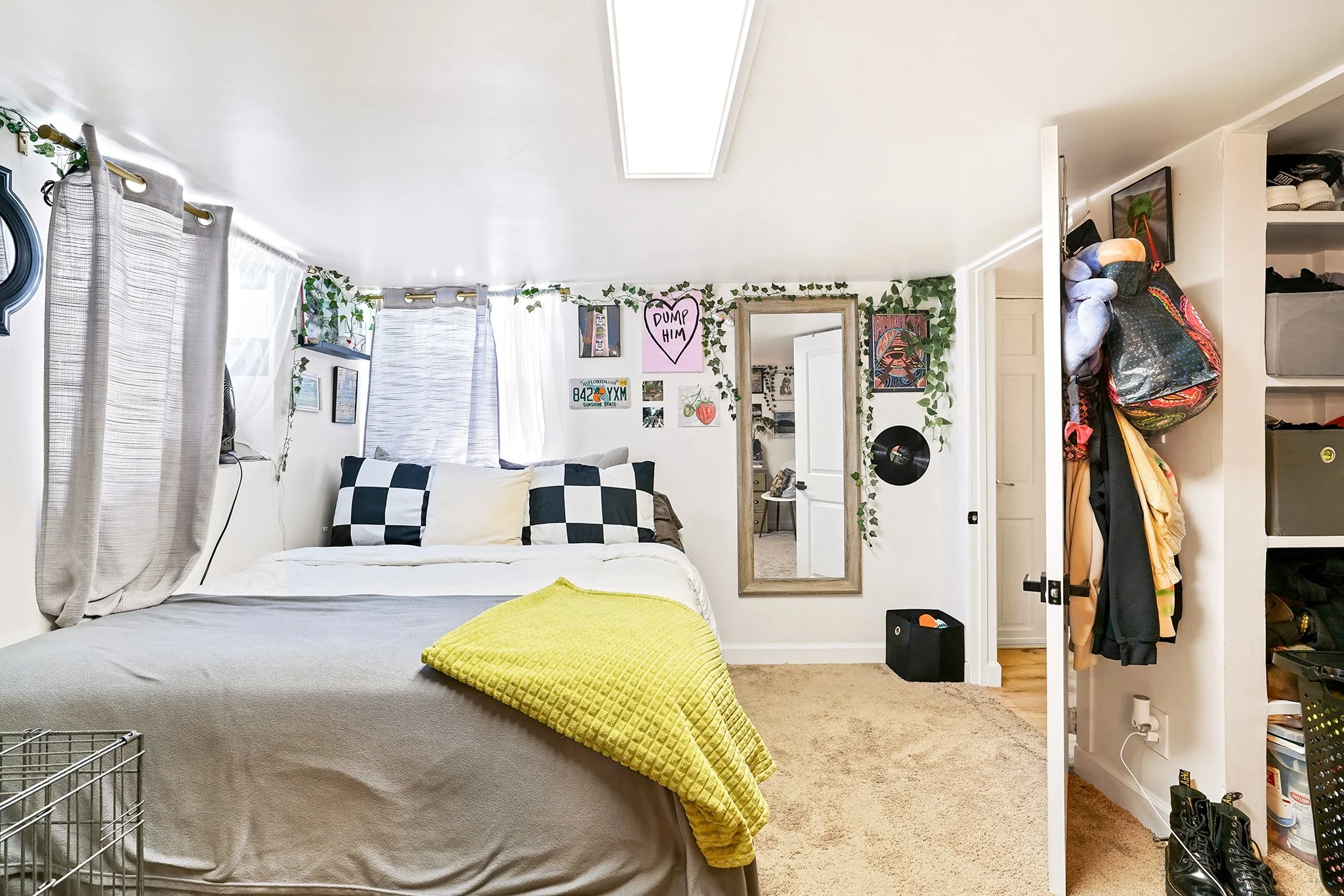 A cozy bedroom with white walls, a bed with black and white checkered pillows and a yellow blanket, and wall decorations including a pink heart saying 'DUMP HIM' and various pictures. There are light-colored curtains covering the window, a large mirr