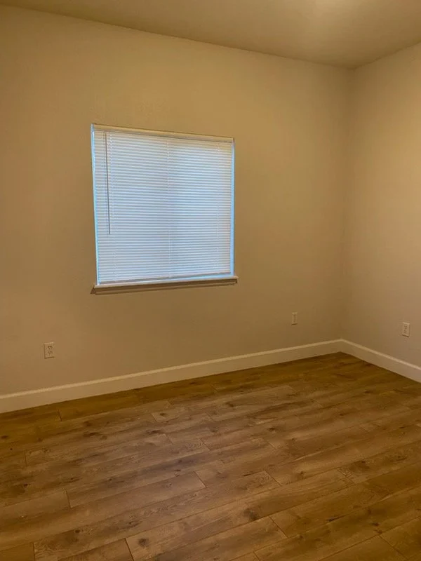 Empty room with a window covered by closed blinds, beige walls, hardwood floor, and white baseboards.
