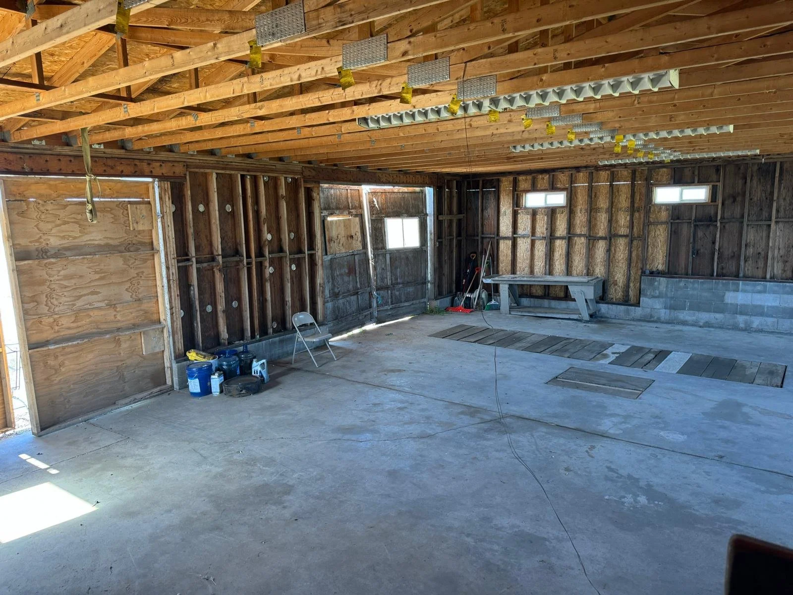 Interior of a garage with unfinished walls, wooden ceiling beams, construction tools, paint cans, a folding chair, and plywood on the floor.