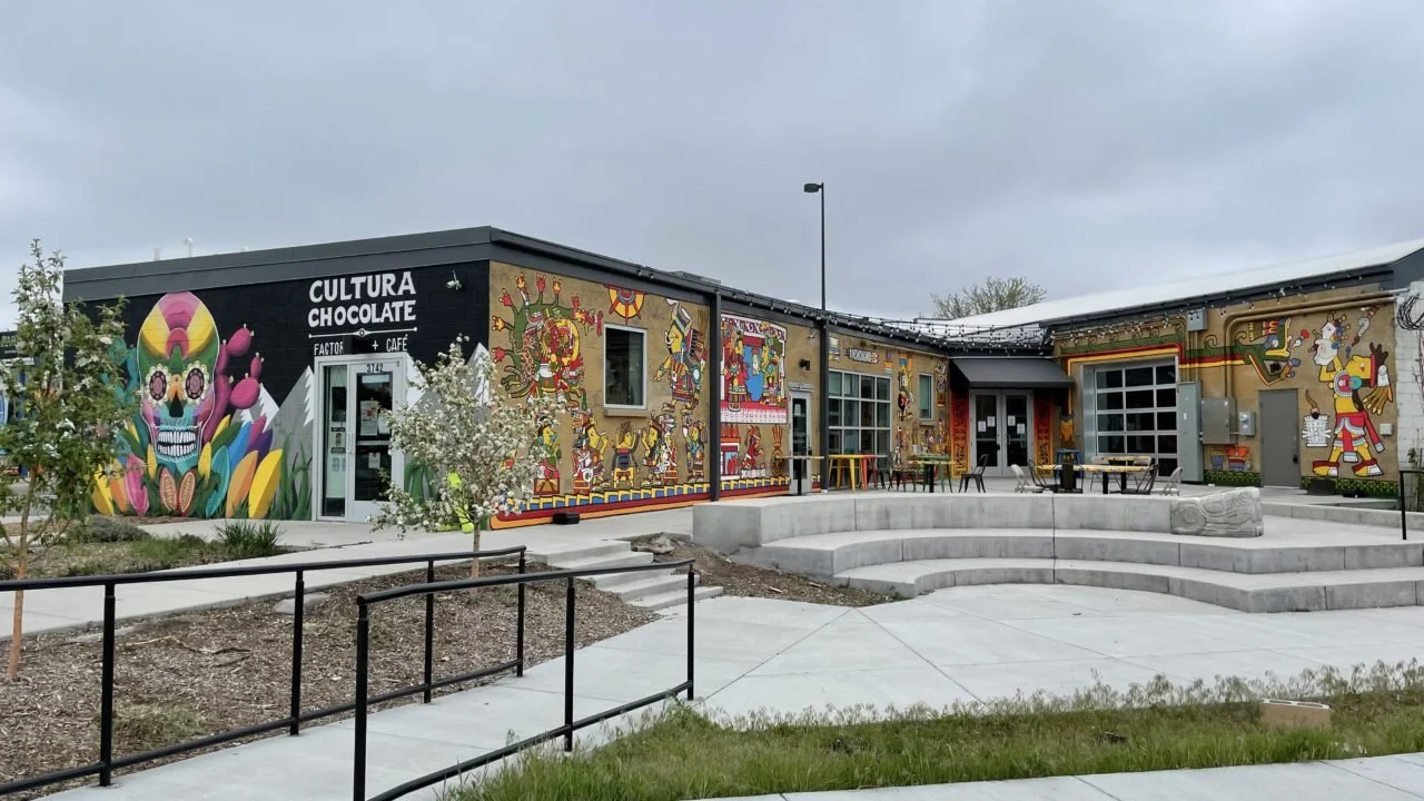 A mural-covered building with a sign that reads 'CULTURA CHOCOLATE' and 'FACTOR CAFE,' featuring colorful artwork including skulls, animals, and abstract designs, with outdoor seating and a concrete stage area.