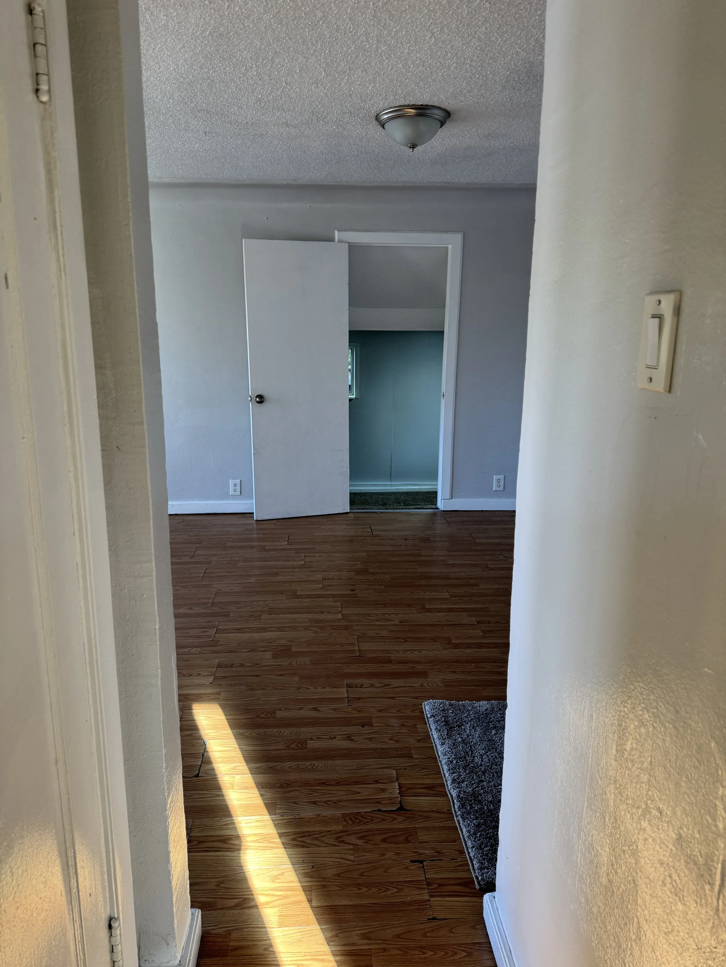 An interior view of a room with wooden flooring, white walls, a closed white door, and a small open door leading to another space with a small window.