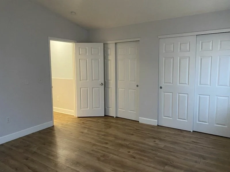 Empty room with light gray walls, wooden flooring, and multiple white closet doors, one of which is partially open, revealing a passage to another room.