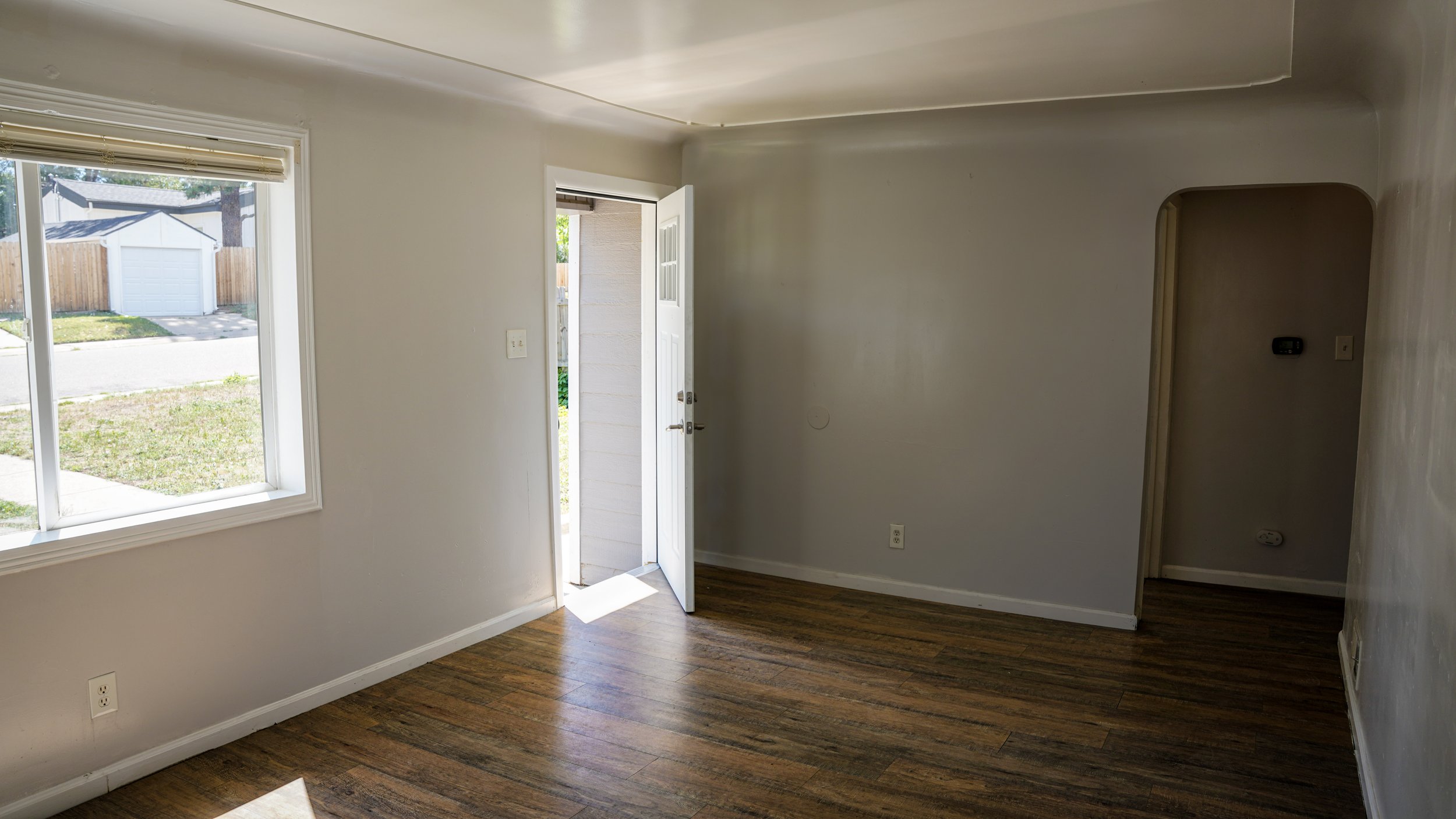 Empty room with a window, open front door letting sunlight in, hardwood floors, and an arched doorway leading to another area.
