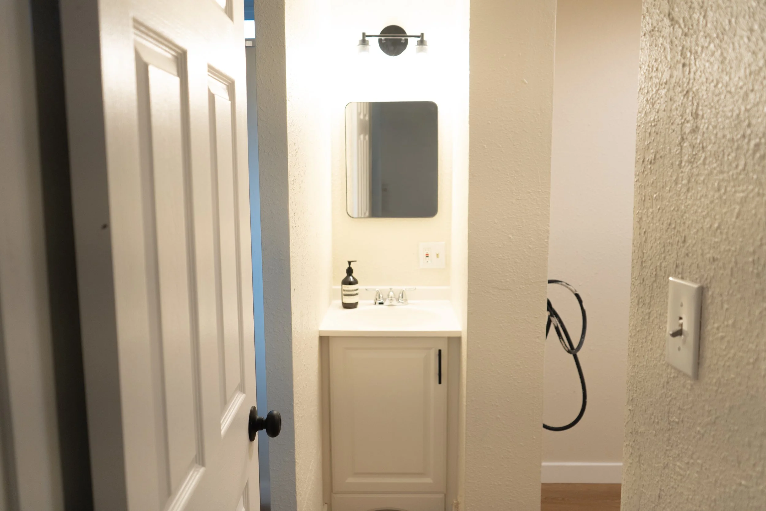 A small bathroom vanity with a white countertop, a black soap dispenser, and a mirror above it, illuminated by a wall-mounted light fixture.