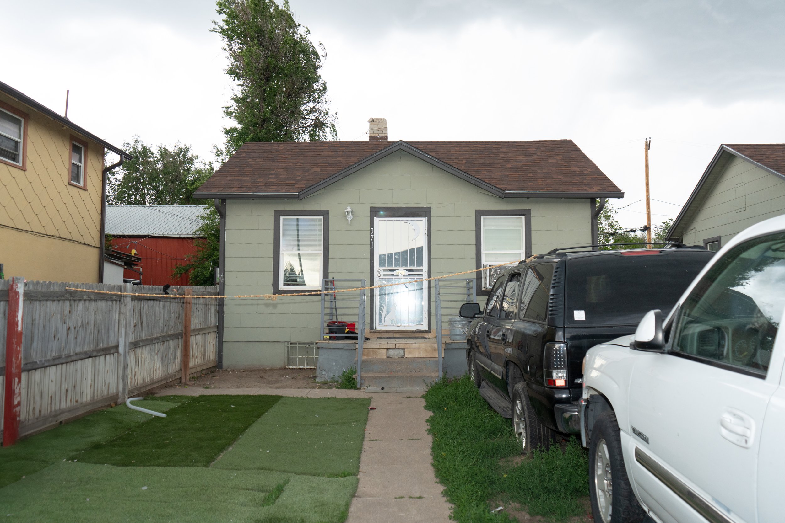 A small house with a green exterior and brown roof, two windows, and a glass door with a moon decal, surrounded by two parked vehicles and a wooden fence, during daytime with an overcast sky.