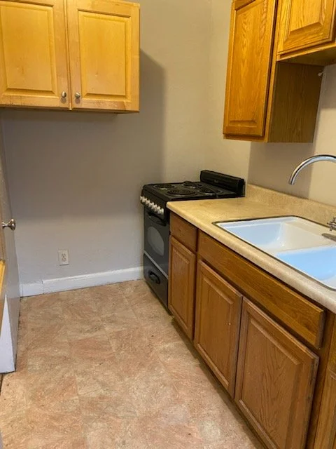Compact kitchen with wooden cabinets, a small stove, a sink, and beige countertops.