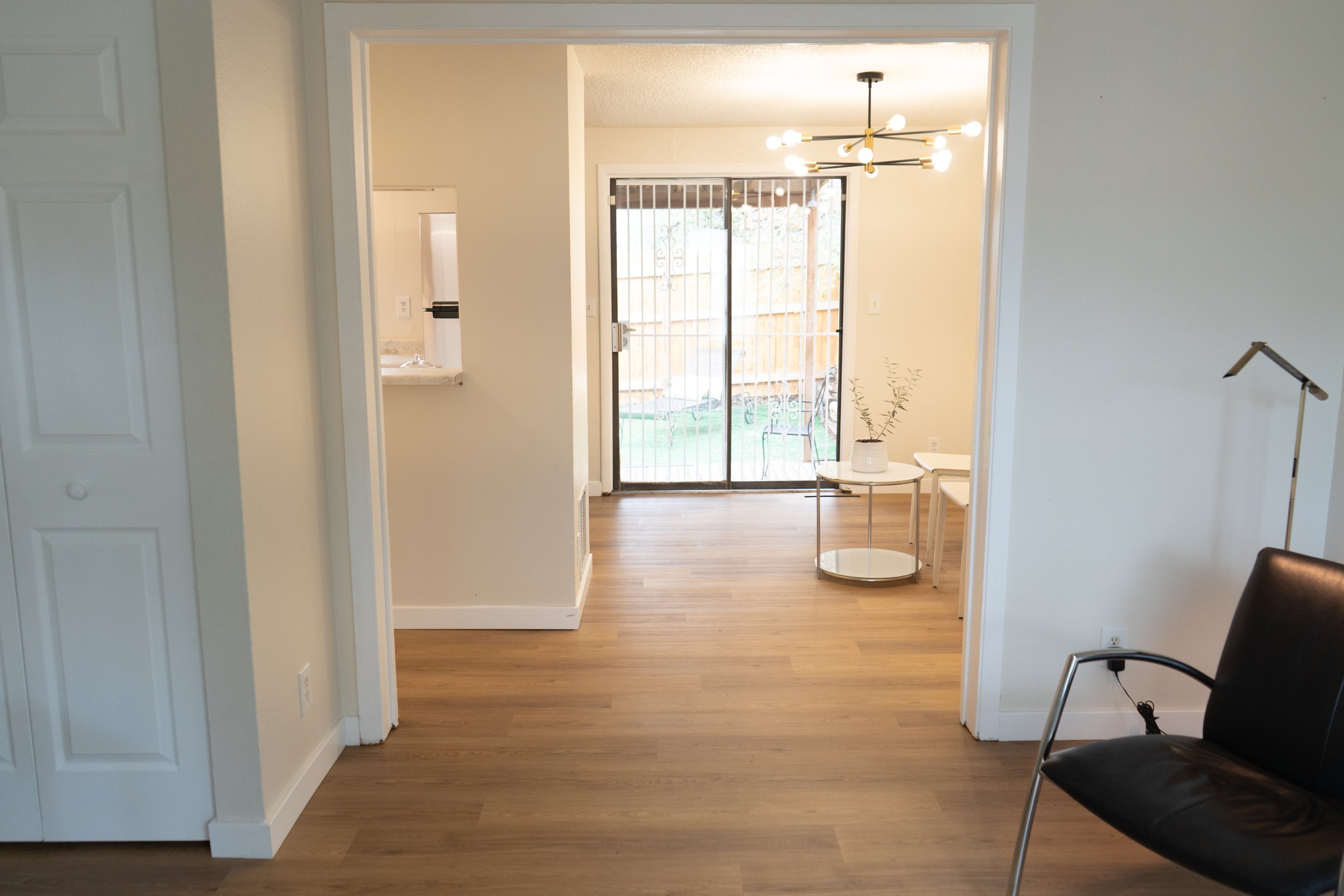 Living room with hardwood floor, sliding glass door leading outside, dining area with modern chandelier, small side table with plant, black chair, and a partially visible wall with white doors.