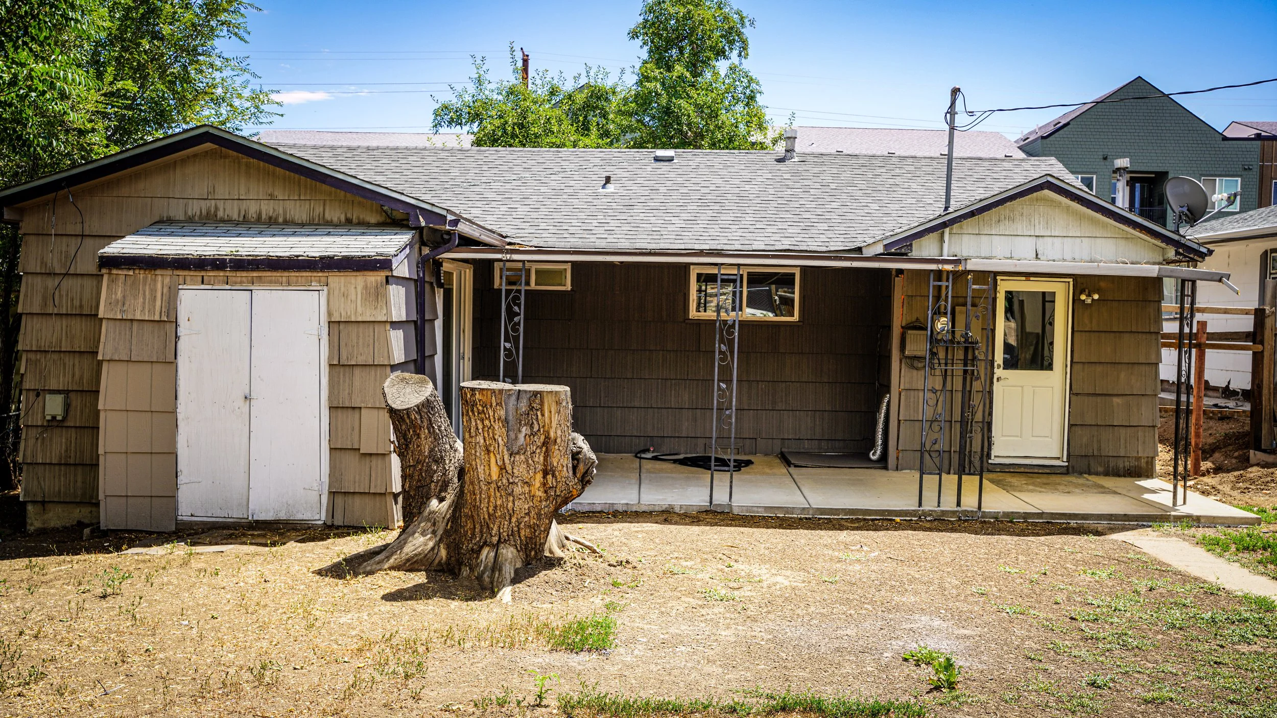 Backyard view of a small, weathered house with brown siding and a gray shingled roof. There is a small porch area with metal supports, a white door, and small windows. A large tree stump and a cut tree trunk are in the yard, which has patchy dirt and