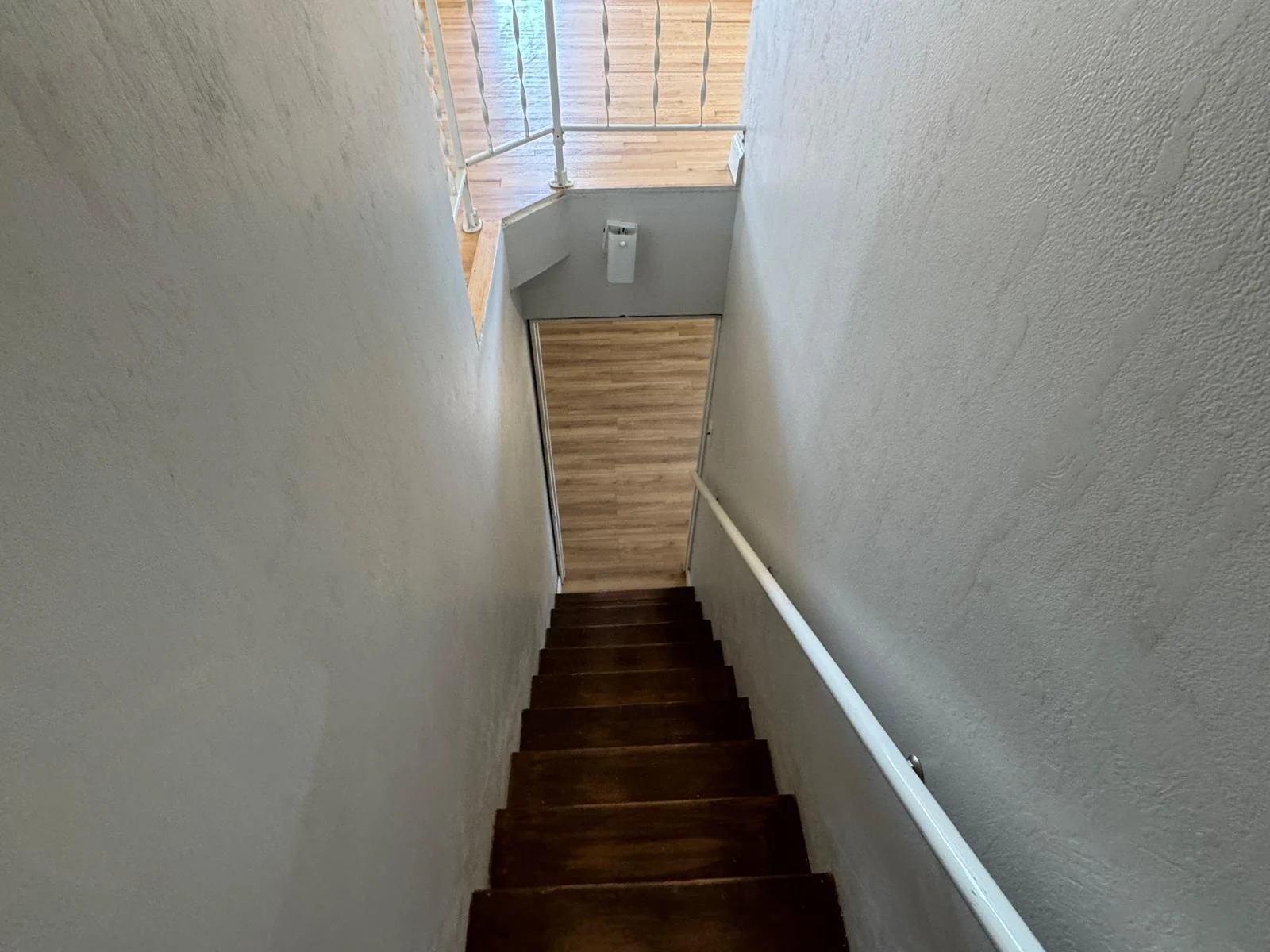 Looking down a staircase with dark wooden steps and a white handrail on the right side, leading to a small landing and a hallway with light-colored wooden flooring and a white wall with a hand sanitizer dispenser.