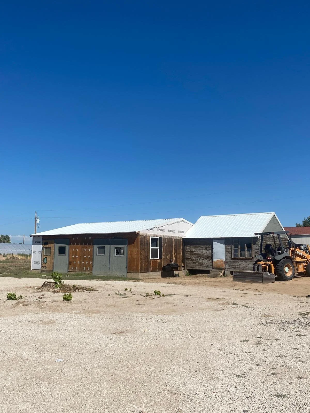 A rural scene with a gravel lot and two weathered buildings, one with a metal roof and the other with a wooden exterior, and a small orange construction vehicle nearby under a clear blue sky.