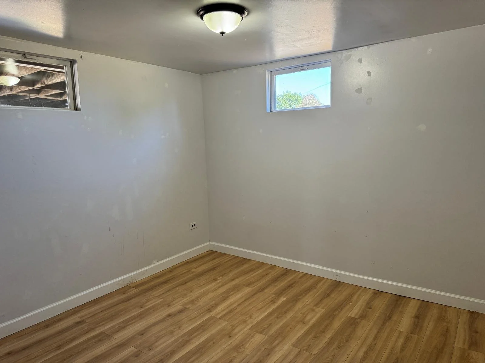 Empty room with white walls, wood laminate flooring, two small rectangular windows near the ceiling, and a flush ceiling light.