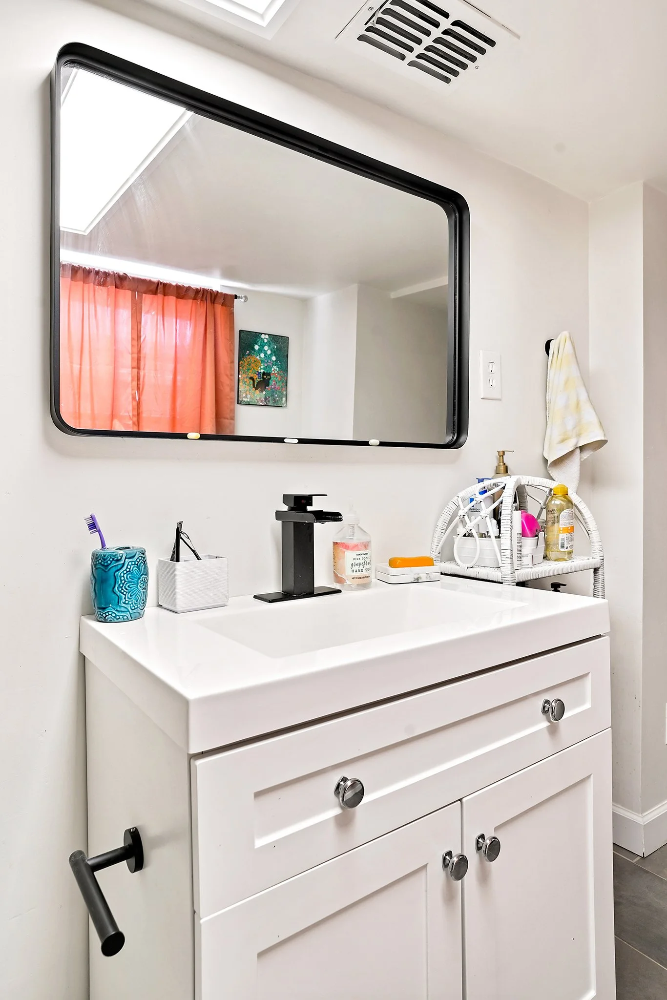 A bathroom vanity with a rectangular mirror, a white sink, a black faucet, and various toiletries. The mirror reflects a window with orange curtains and a colorful artwork on the wall.