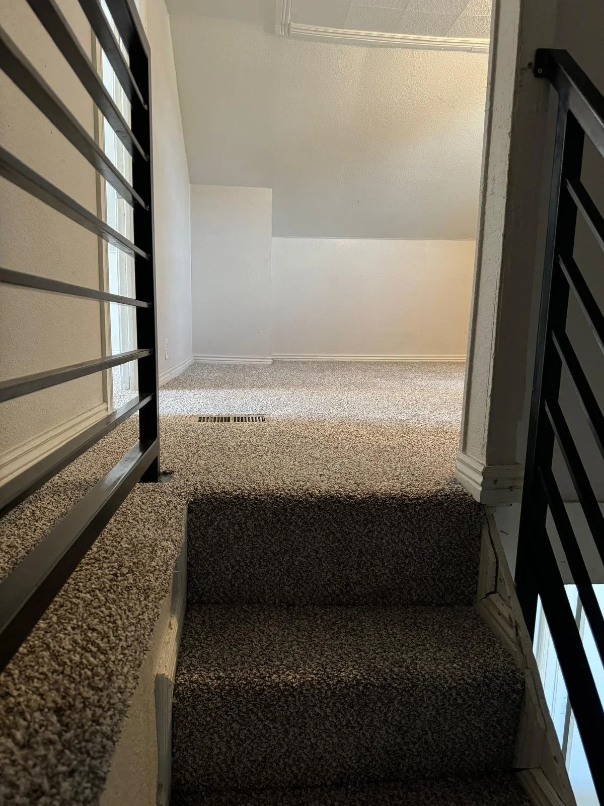 Interior view of a carpeted staircase leading up to a landing with a beige carpet, railing on the sides, and a wall with wood paneling and a floor vent.