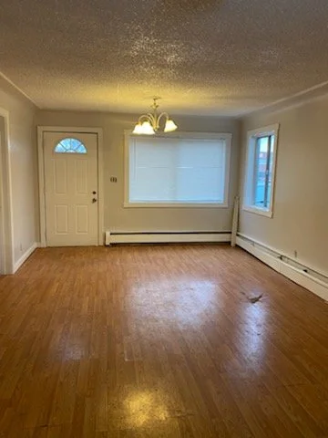 Empty living room with hardwood floors, a front door, and two windows, one with blinds and the other clear, in a house during daytime.