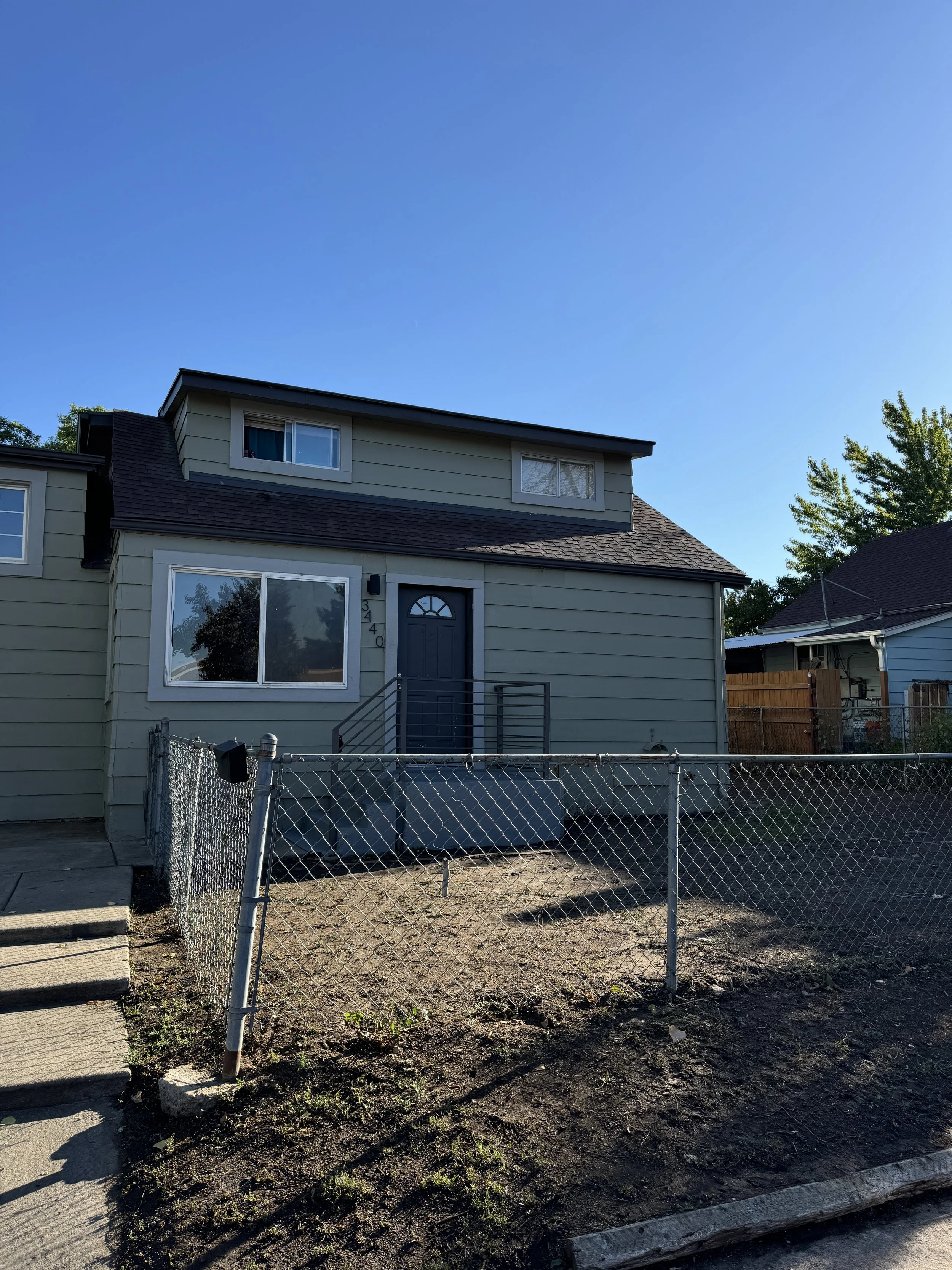 A two-story house with light gray siding and dark gray roof, featuring a front door with steps, a large window, and a smaller window on the upper floor. There is a chain-link fence in the front yard with some dirt and plants, under a clear blue sky.