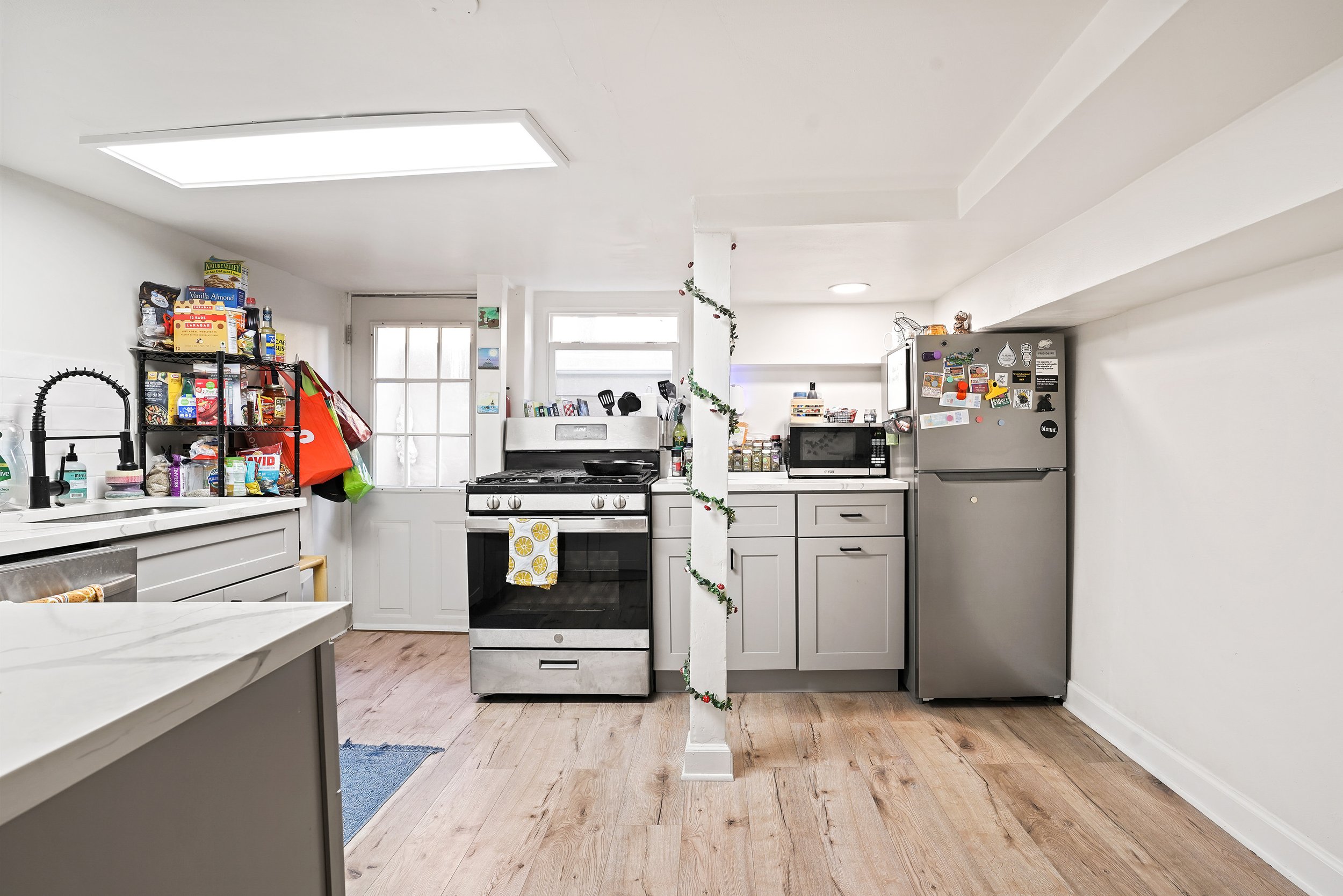 A kitchen with white cabinets, a gray refrigerator with magnets, a stove with a lemon-themed towel, and a black microwave. There is a shelf with food on the left, a window and door at the back, and a careful decoration with a garland on a column in t