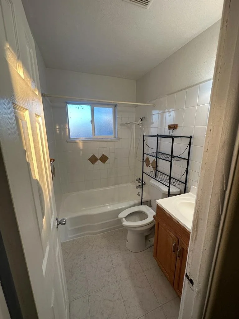 Small bathroom with a bathtub, frosted window, toilet, wooden cabinet with a sink, and a black metal shelving unit.