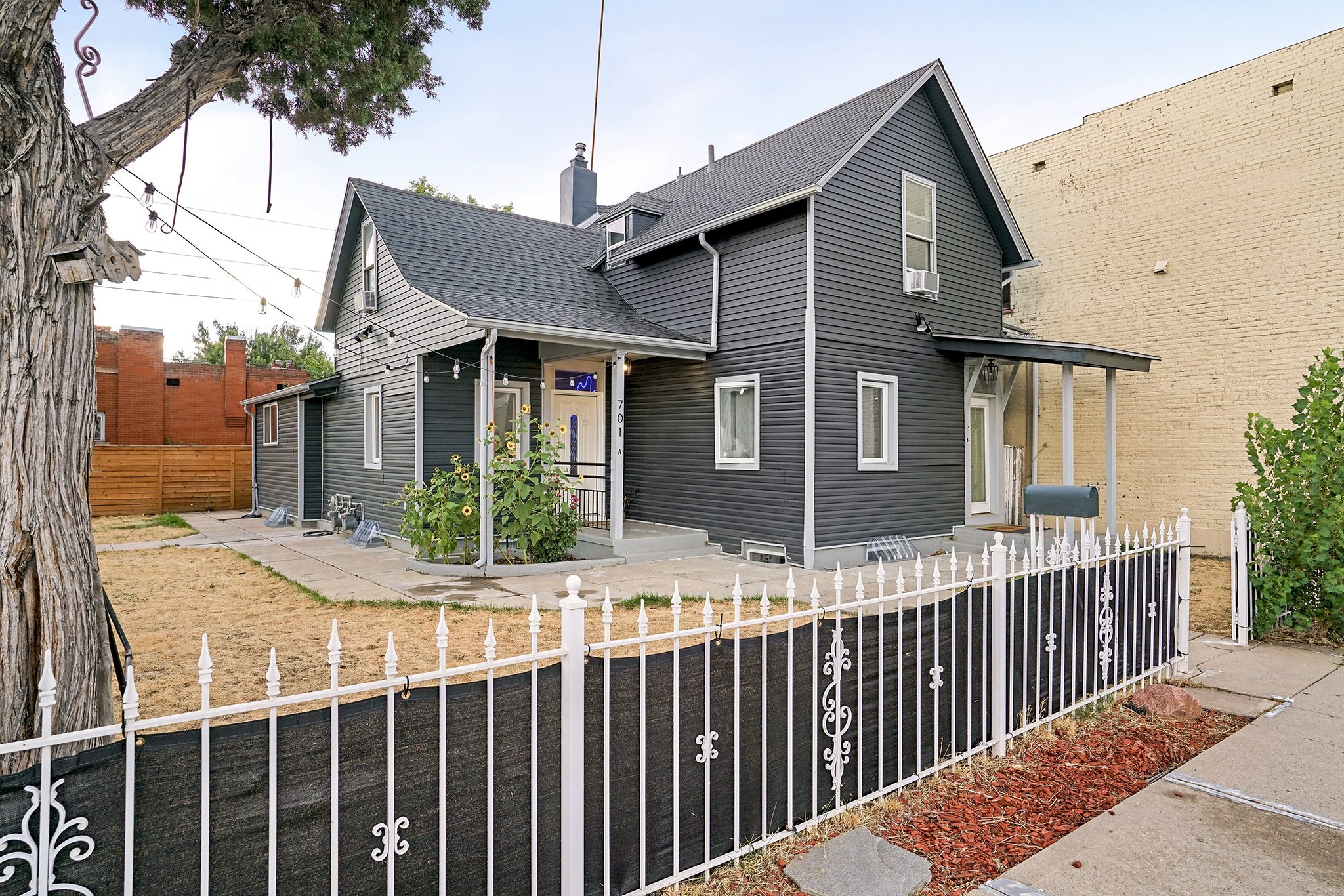 A two-story house with dark gray siding, a gabled roof, and white trim. The front yard has a white picket fence, a small garden with sunflowers, and a paved walkway leading to the front door. String lights are hanging above the yard, and there is a l